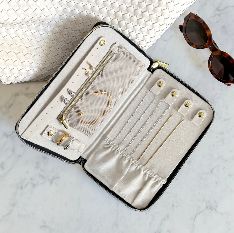 The Jewelry Travel Case in black, shown open on a marble surface, its interior neatly organizing a collection of gold and silver jewelry including rings, earrings, necklaces, and a bracelet.