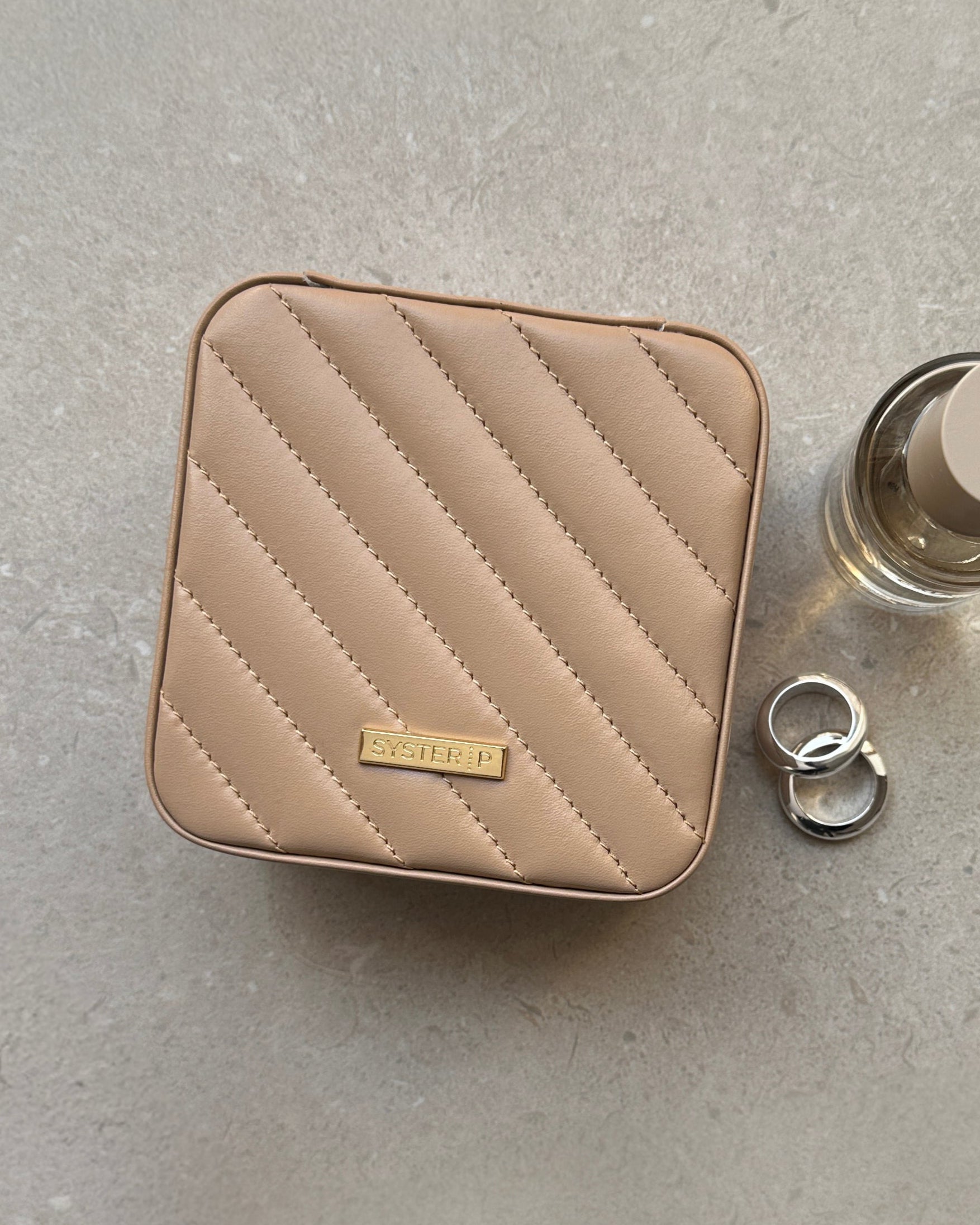 A flat lay of the Syster P Jewelry Case Small in beige, highlighting its quilted vegan leather exterior, styled with two chunky silver rings and a perfume bottle.