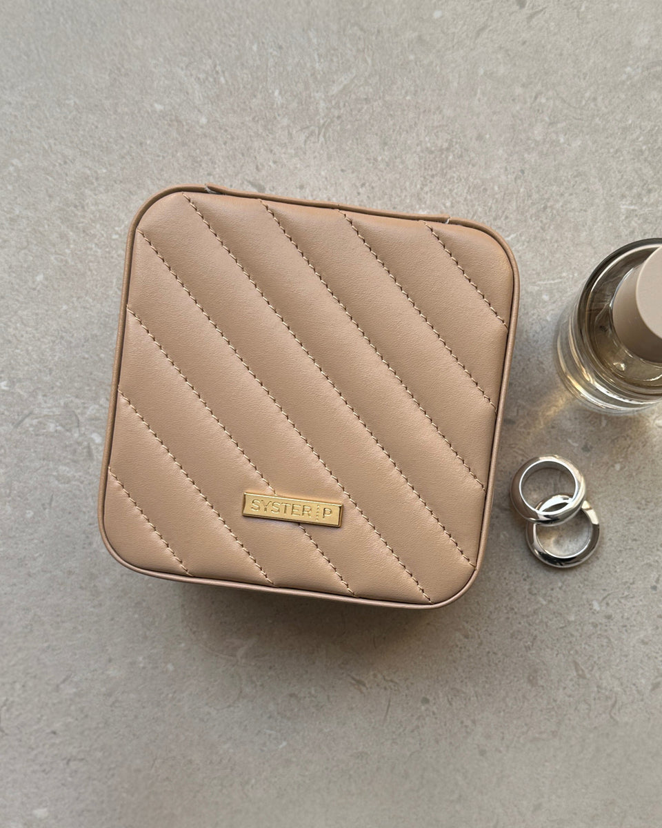 A flat lay of the Syster P Jewelry Case Small in beige, highlighting its quilted vegan leather exterior, styled with two chunky silver rings and a perfume bottle.