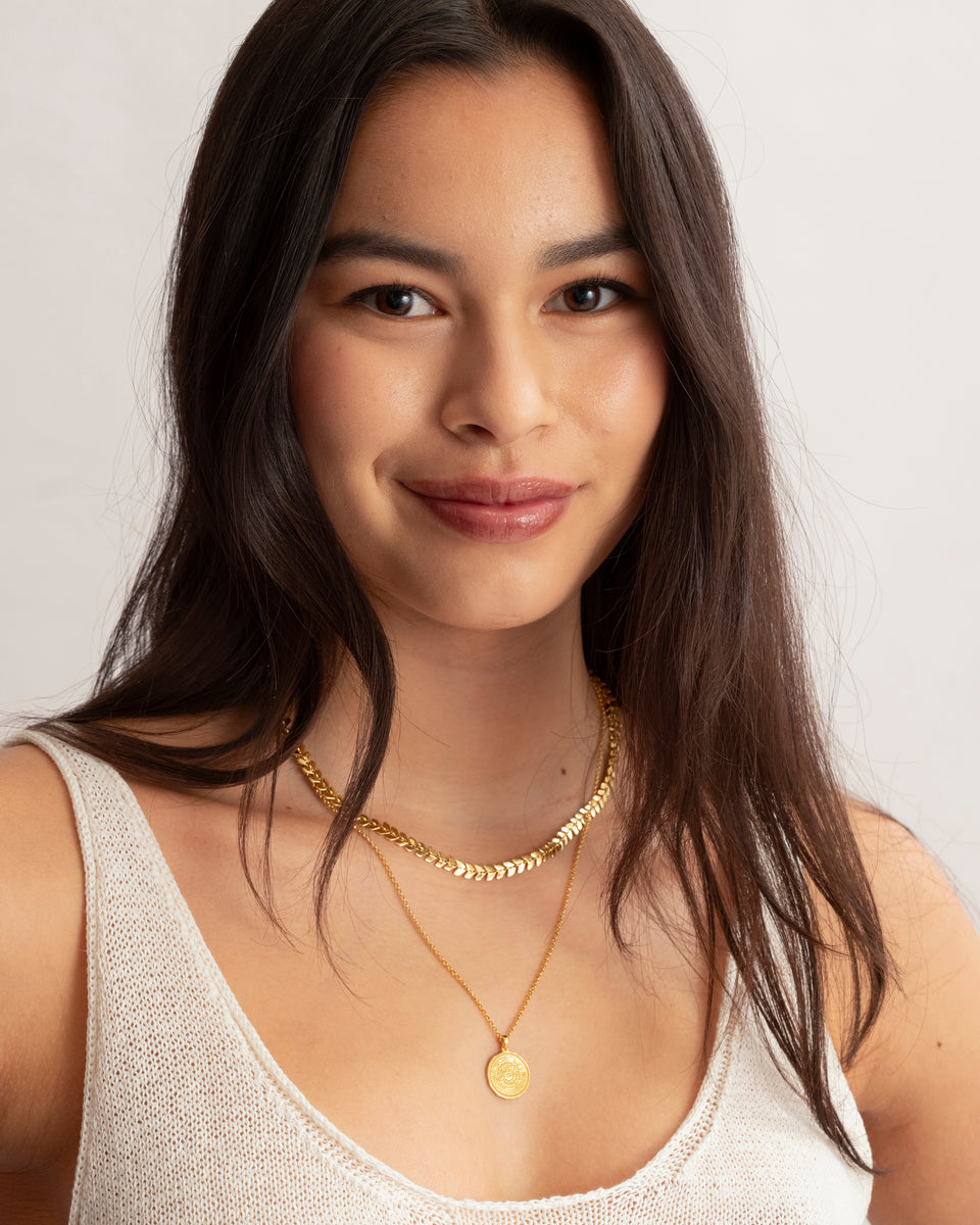 A close-up of a model wearing the Lucky Coin Navigate Necklace in gold, featuring a compass pendant, layered with a thicker gold chain necklace.