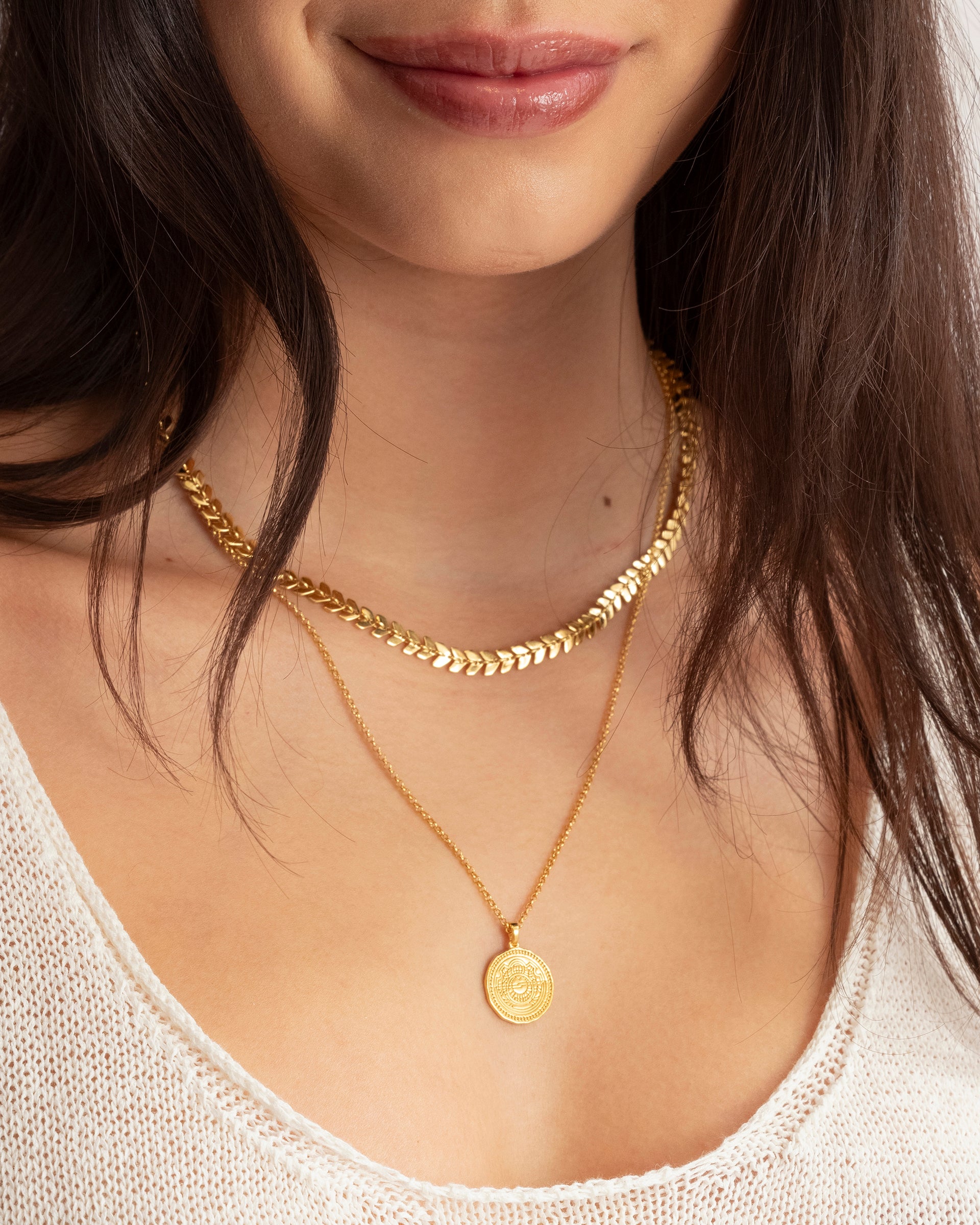 A close-up of a model wearing the Lucky Coin Navigate Necklace in gold, featuring a round compass pendant, layered with a thicker gold necklace with a leaf-like pattern.