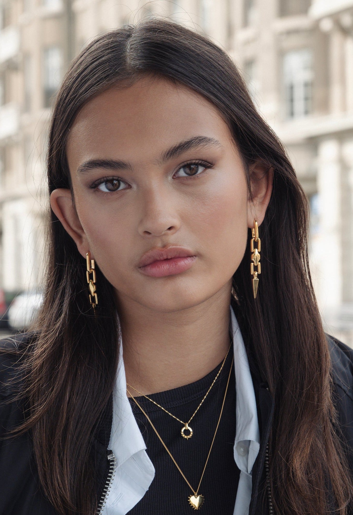 A model wears a pair of gold Marion Long Single Earrings with a spike design, styled with two layered gold necklaces: the Uma Circle Necklace and the True Love Spike Heart Necklace.