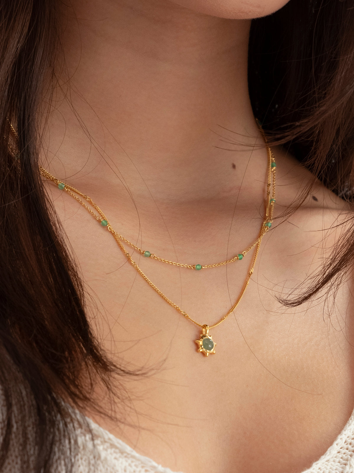 A close-up of a model wearing the Santa Monica Necklace and the Florentina Necklace layered together. The Santa Monica is a delicate gold chain with green onyx beads, and the Florentina features a gold sun pendant with a green jade stone.