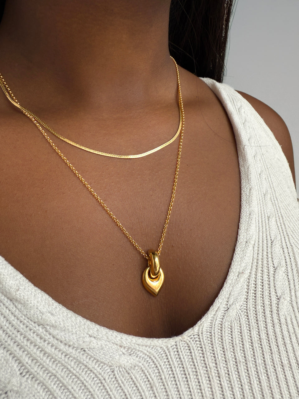 A model wears the Melinda Long Necklace in gold, featuring a chunky leaf-shaped pendant, layered with a shorter gold herringbone chain.