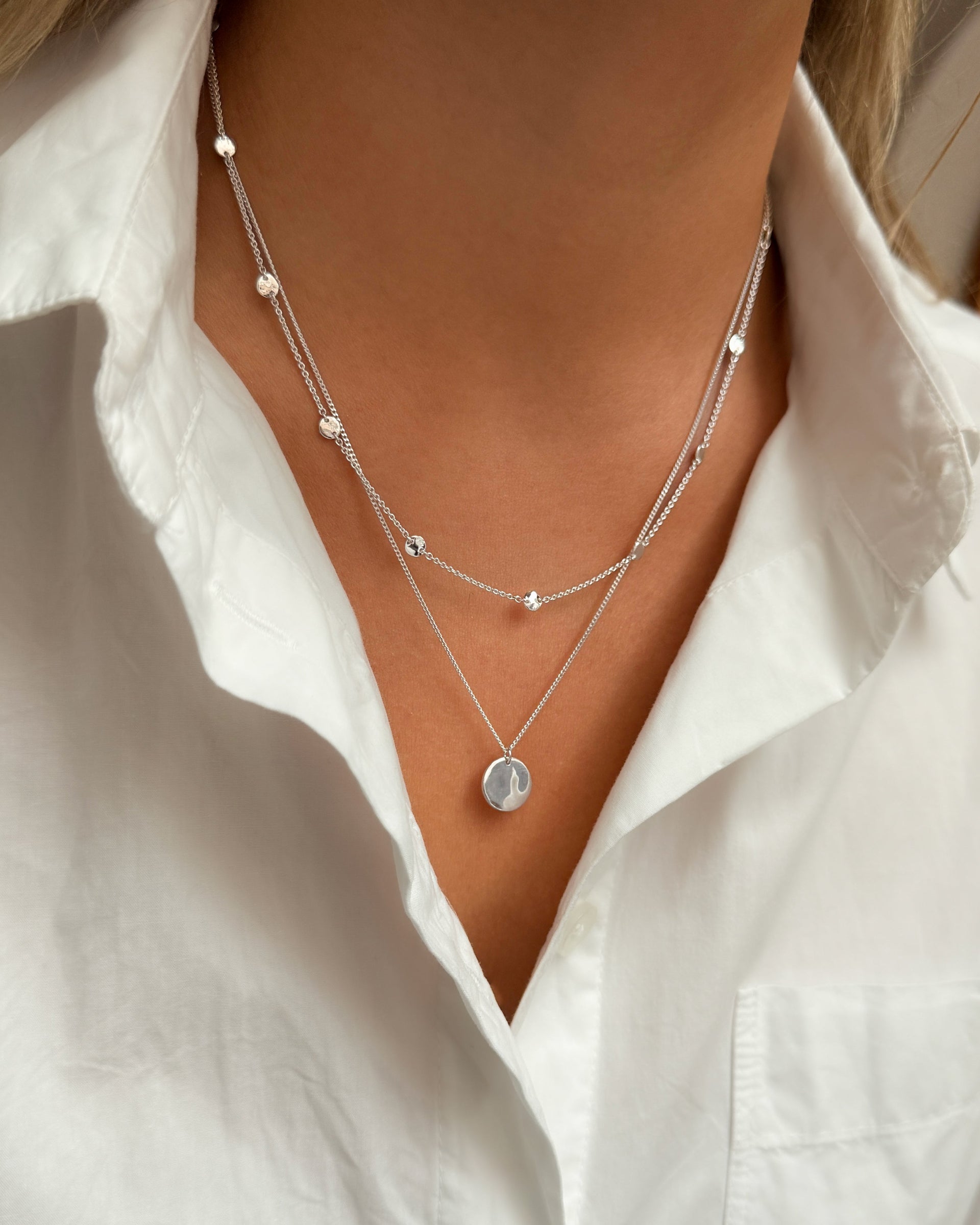 A close-up of a model wearing the Solea Necklace Silver, which has a delicate chain adorned with small hammered silver circles, layered with another silver necklace featuring a round hammered pendant.
