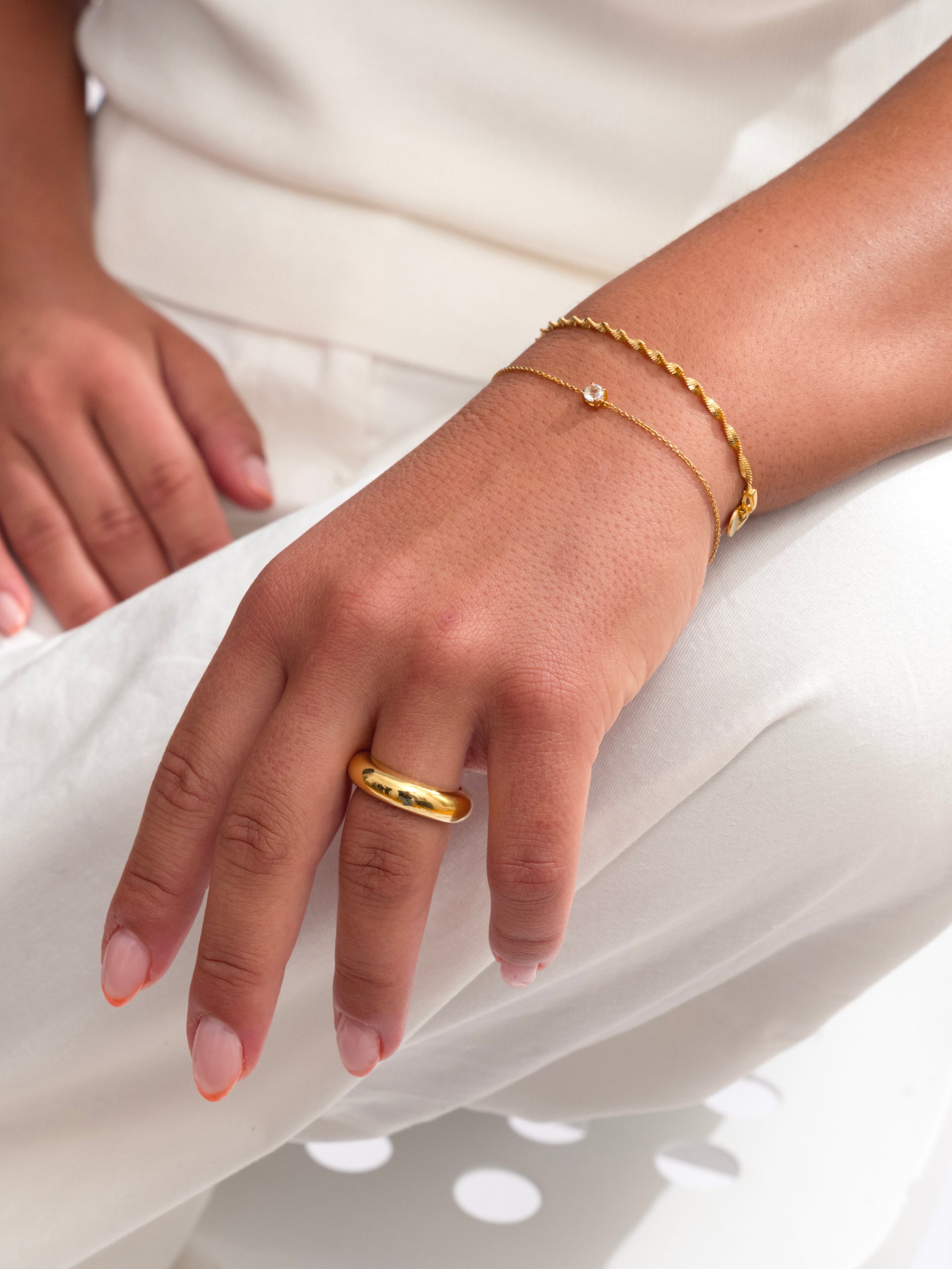 A close-up of a model wearing the gold Minimalistica Solo Nova Bracelet, featuring a delicate chain with a small white topaz. She has layered it with a twisted gold bracelet and is also wearing a bold gold ring on her finger.