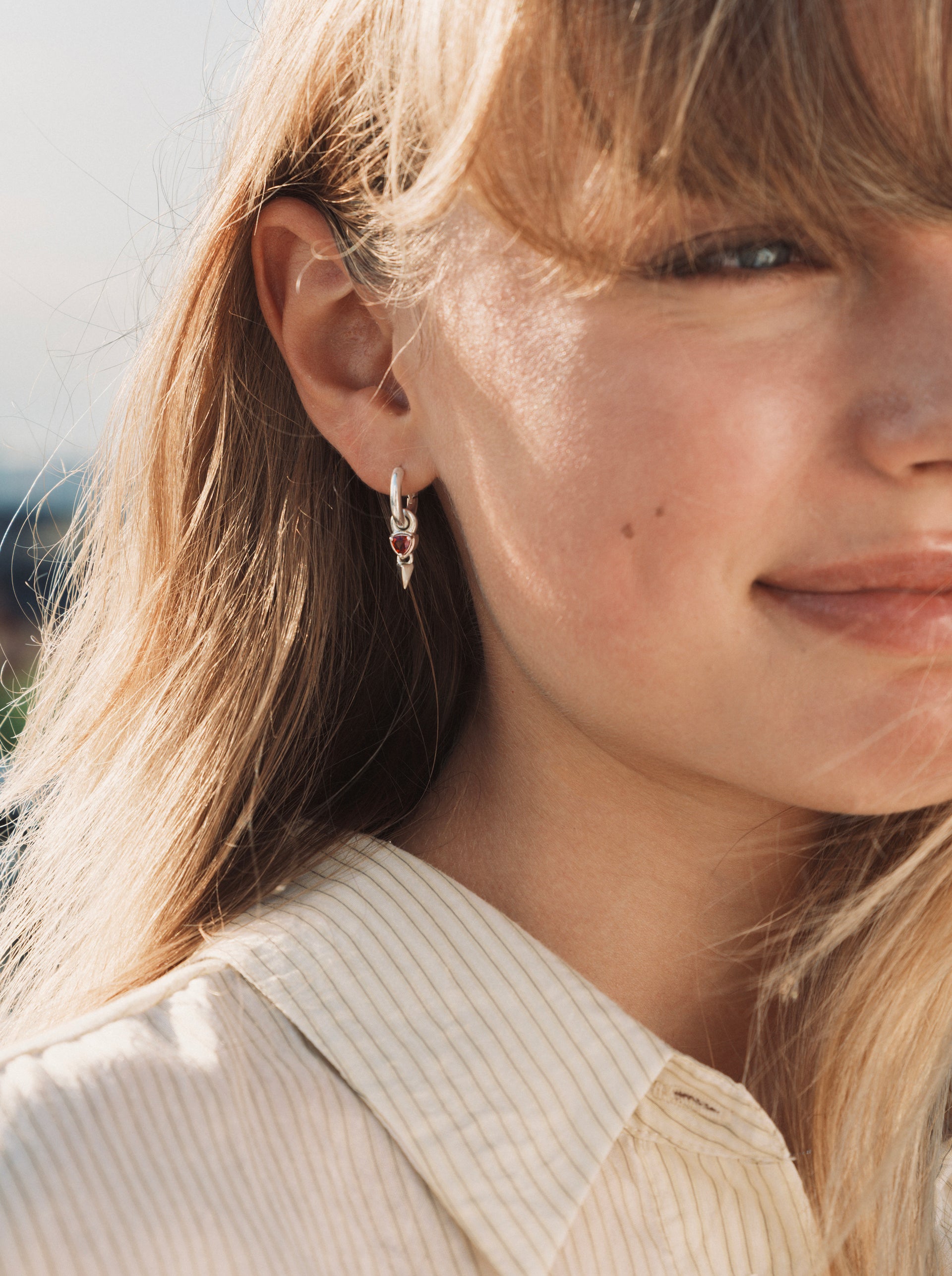 A close-up shot of a model wearing the Mrs H Spike Earrings in silver, highlighting the chunky hoop with its dangling charm featuring a red garnet stone and a silver spike.