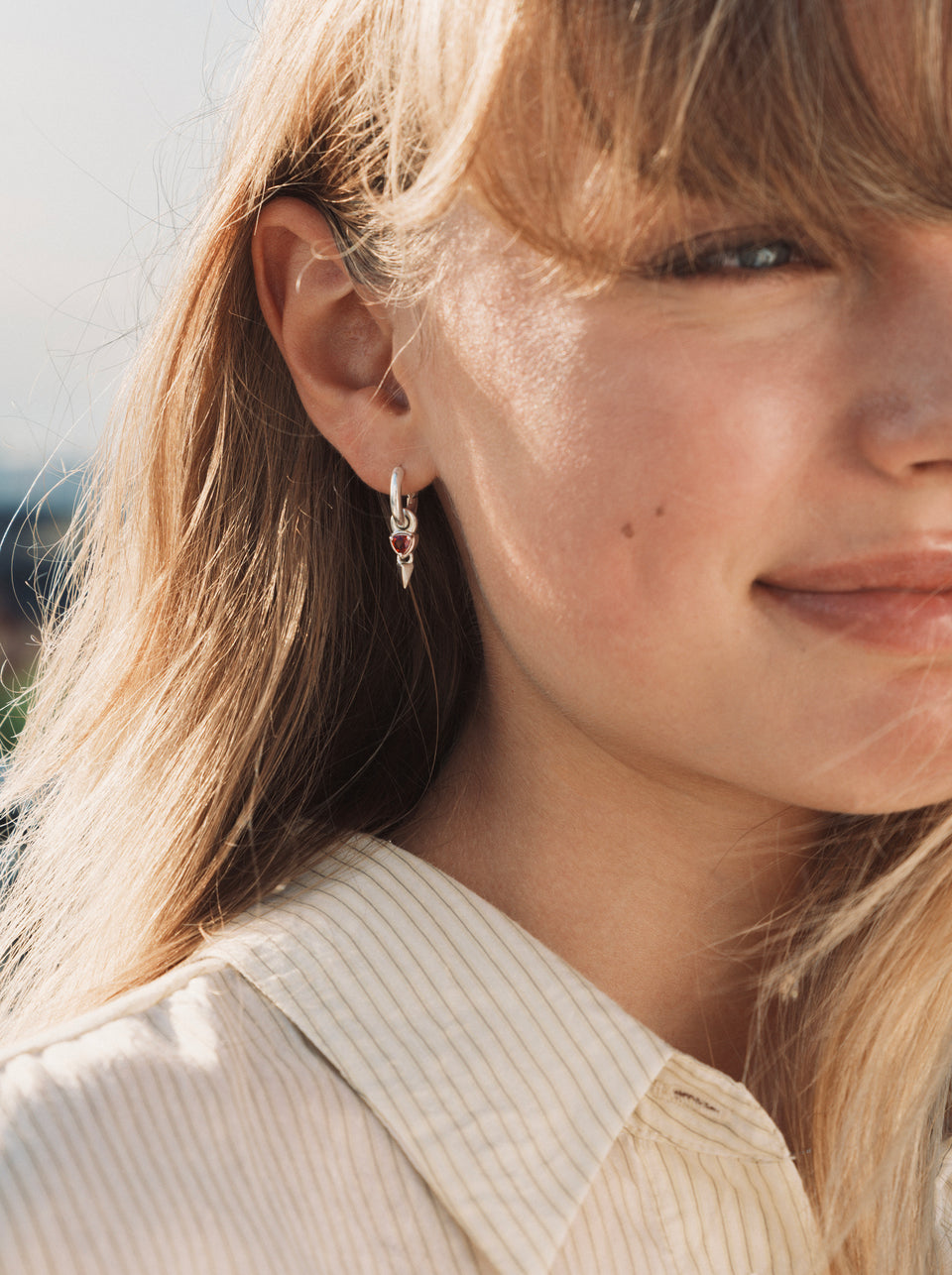A close-up shot of a model wearing the Mrs H Spike Earrings in silver, highlighting the chunky hoop with its dangling charm featuring a red garnet stone and a silver spike.