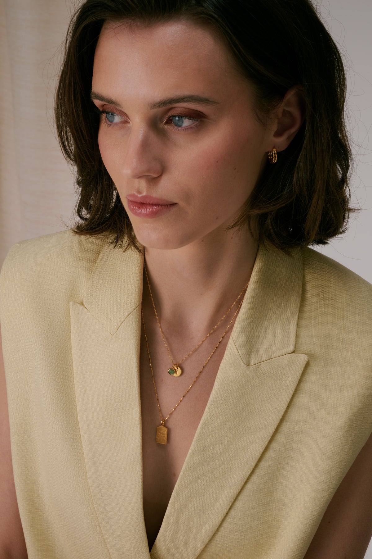 Two gold necklaces layered - short chain with letter pendant and longer oval chain, worn with textured hoop earrings.