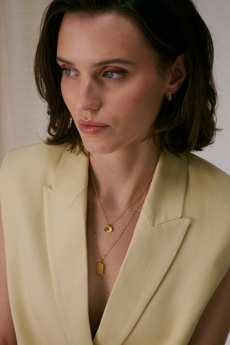 Layered gold necklace set with Mini Letter E pendant and green heart charm on top chain, rectangular pendant on bottom chain, paired with small textured gold hoop earrings.