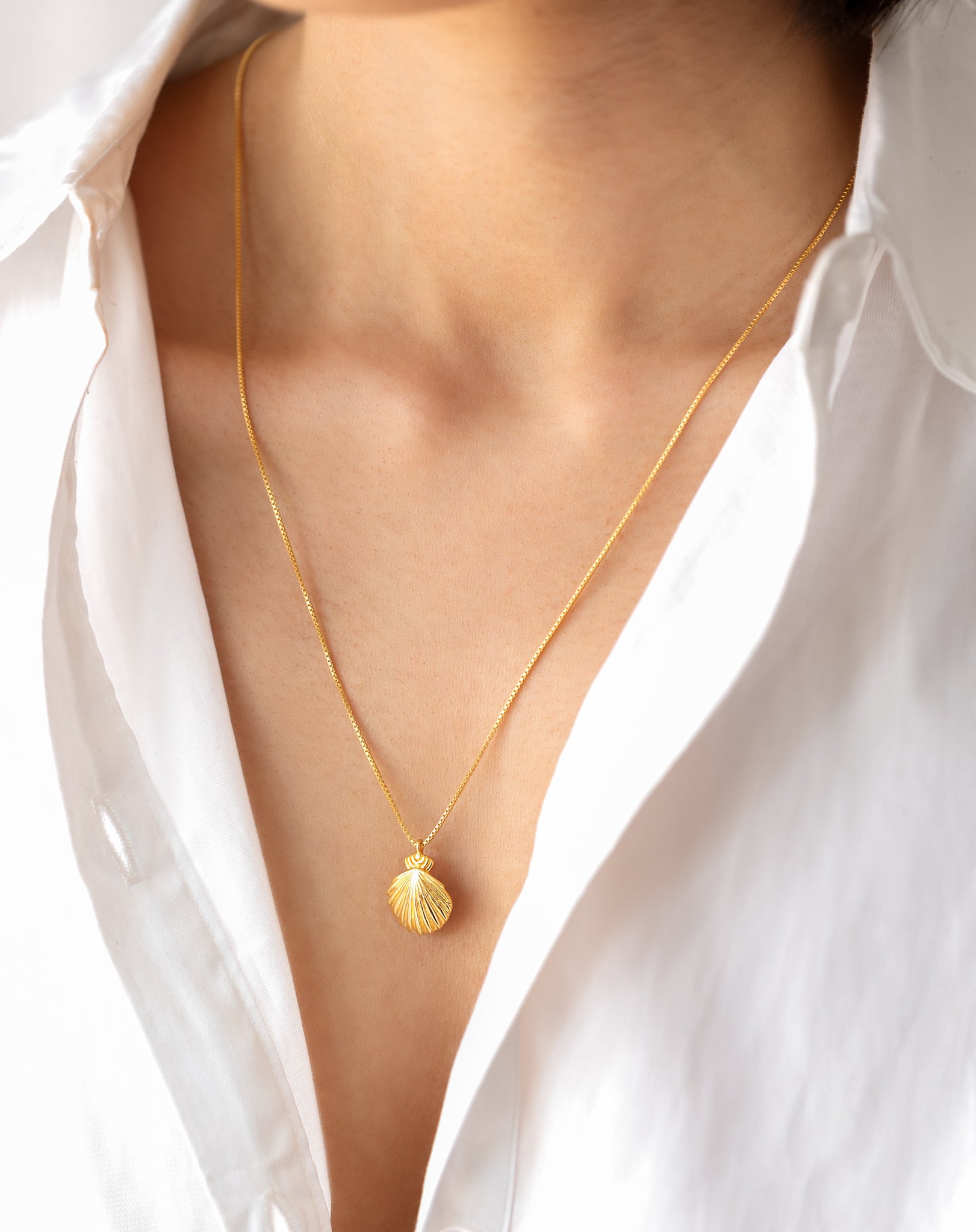 A model wears the gold Beaches Shell Necklace, its delicate shell pendant resting on her collarbone, styled with a crisp white shirt.