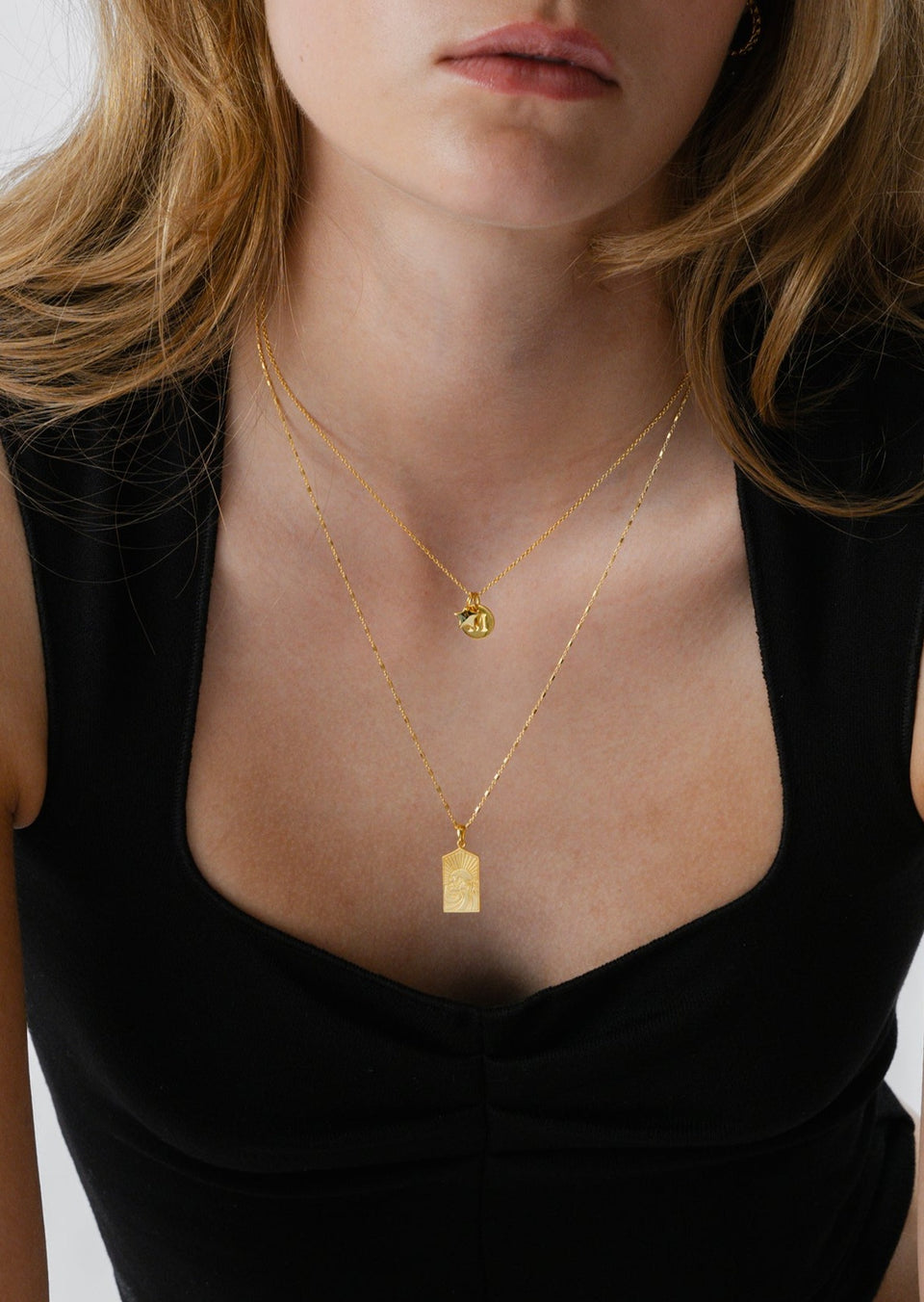 A close-up shot of a model wearing two layered gold necklaces. The shorter chain holds a small, round 'M' initial pendant, while the longer chain features a rectangular pendant with an engraved sun and wave design.