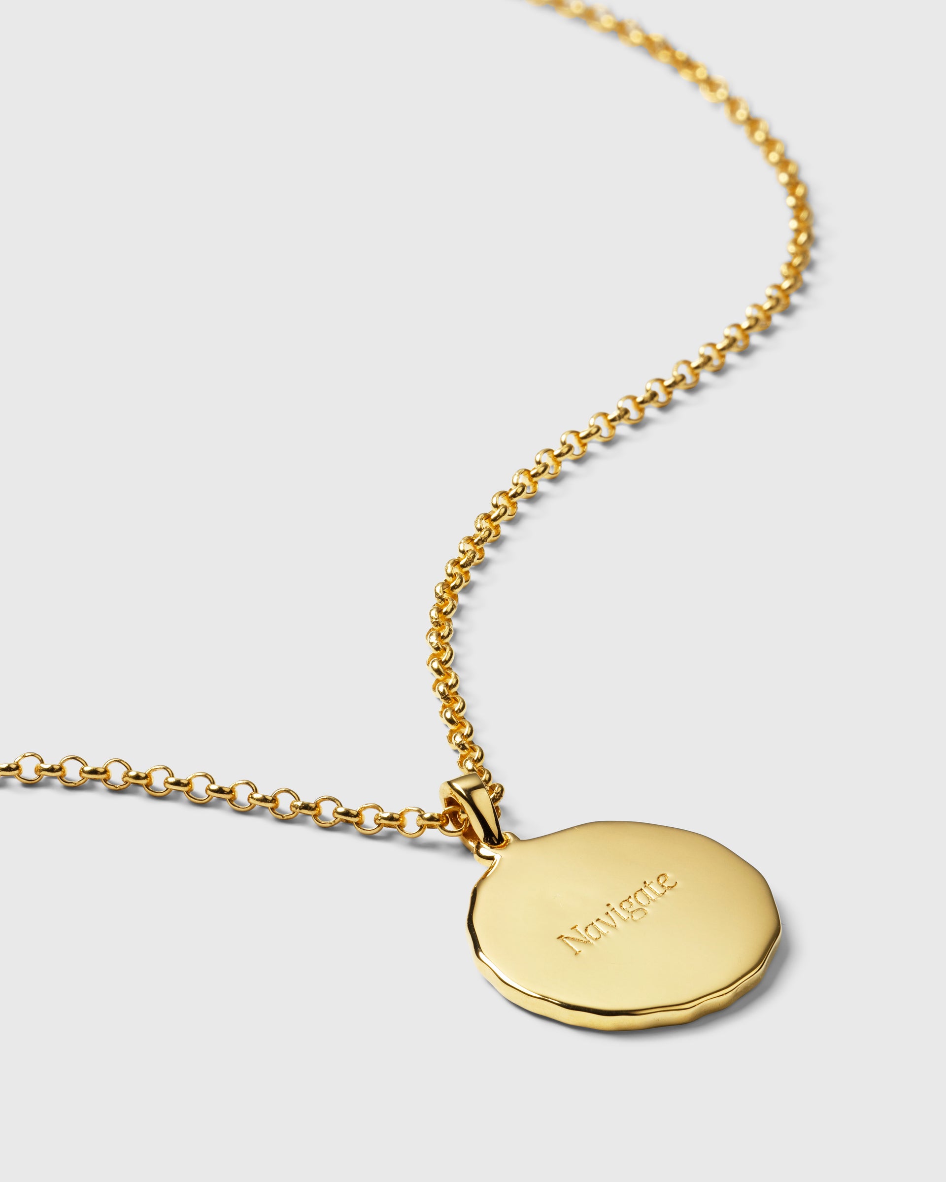 A product shot of the Lucky Coin Navigate Necklace in gold, showing the back of the round pendant engraved with the word 'Navigate'.