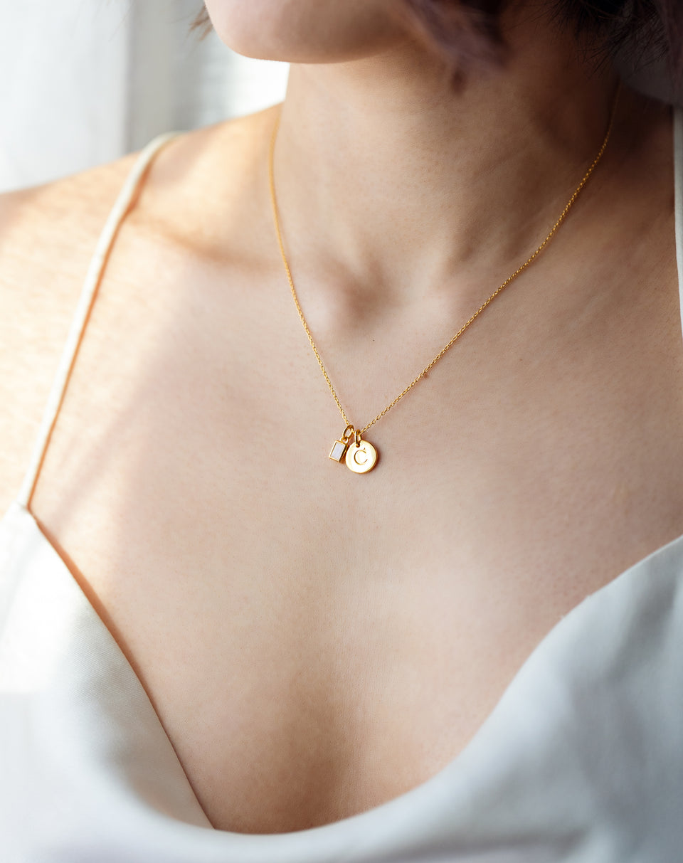 A close-up of a model wearing the gold Beloved Squared Pendant with a moonstone, styled on a fine gold chain alongside a round gold letter 'C' pendant.