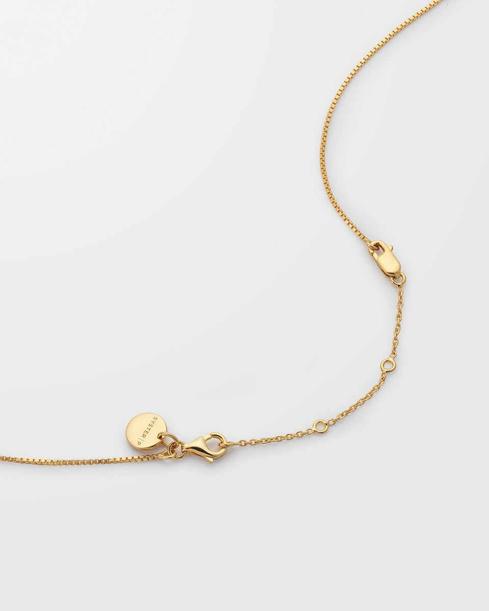 A product shot of the Syster P Extension Chain in gold, demonstrating how it attaches to another necklace to add length, with a close-up on the lobster clasps and adjustable rings.