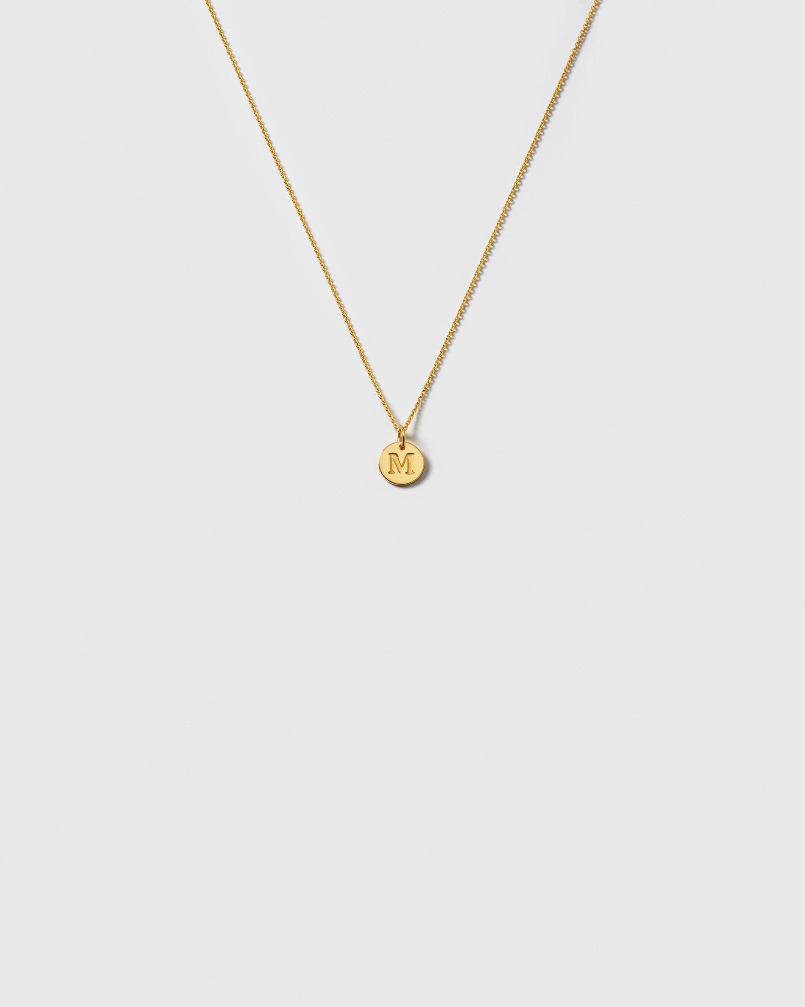 A product shot of the Beloved Baby Chain Gold, a fine anchor chain necklace, styled with a small, round gold pendant engraved with the initial 'M'.