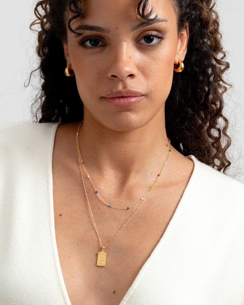 A close-up of a model wearing the Santa Monica Necklace in gold, a fine chain with small colorful gemstones, layered with another gold necklace featuring a rectangular pendant and paired with chunky gold hoop earrings.