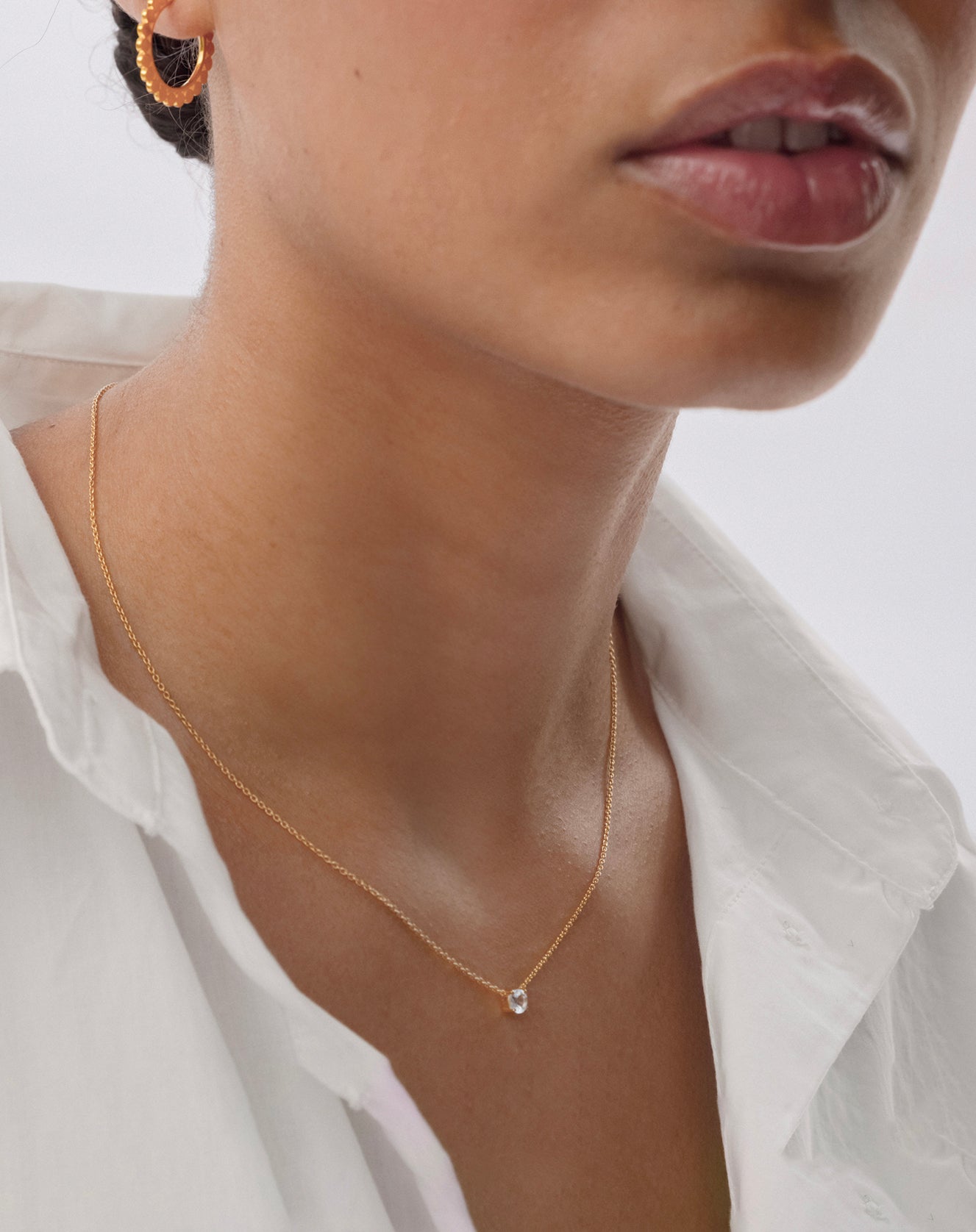 A close-up shot of a model wearing the Minimalistica Solo Nova Necklace in gold, featuring a delicate chain with a single white topaz stone, paired with a textured gold hoop earring.