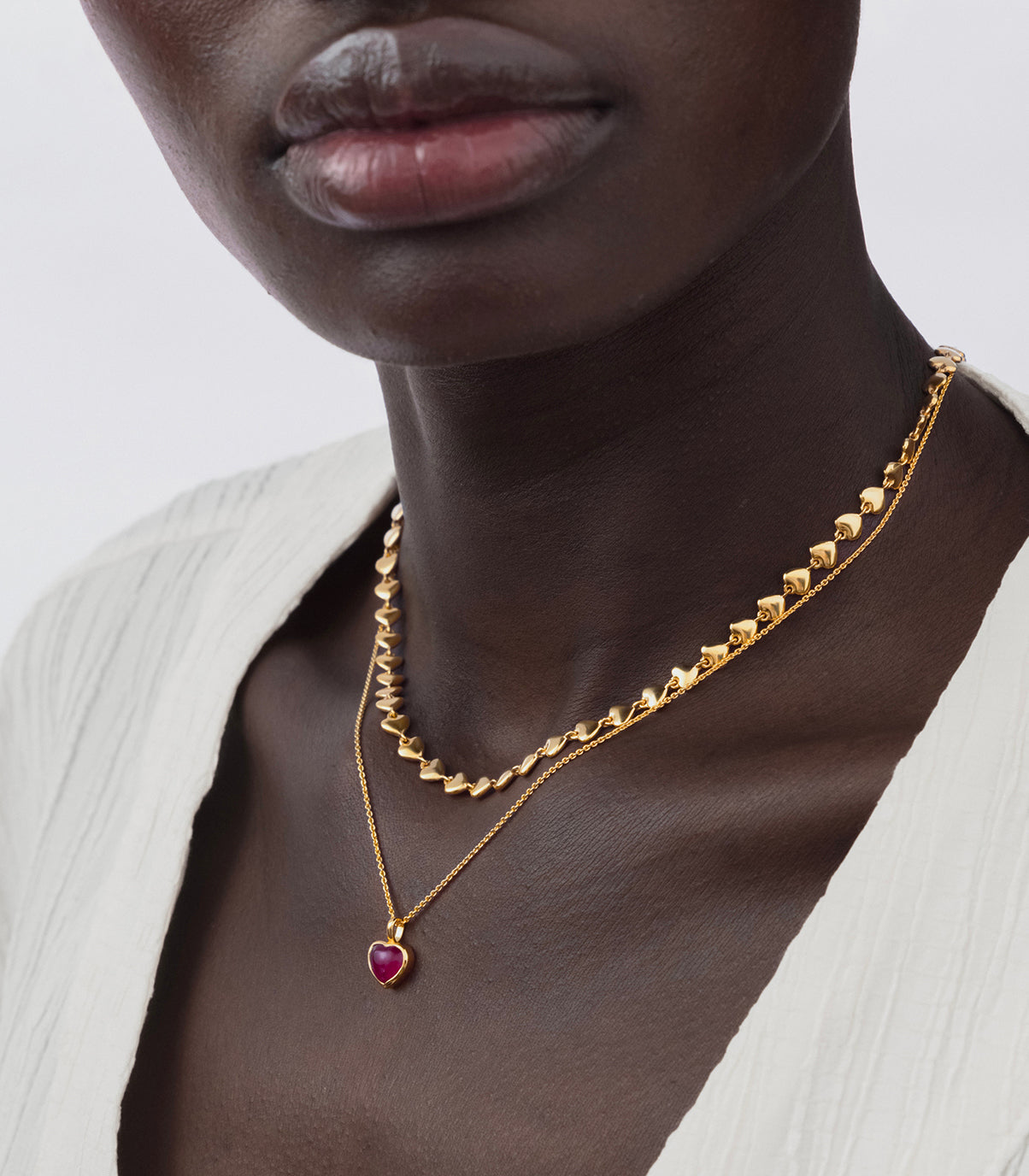 A model wears the gold La La Love Necklace with a pink quartz heart pendant, layered with another Syster P gold necklace composed of small, linked hearts.