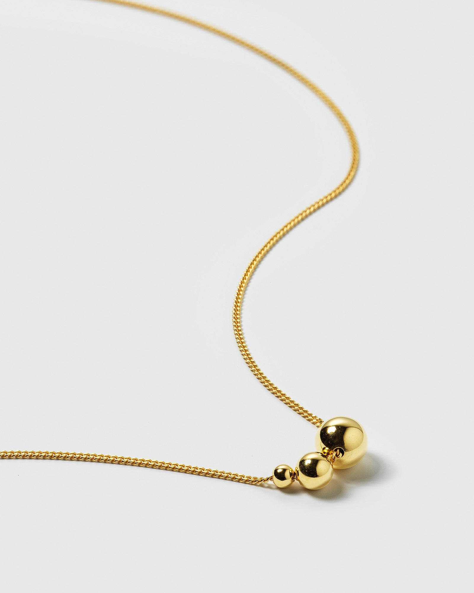 A close-up product shot of the Universe Necklace in gold, showing the fine chain and the three polished gold balls of varying sizes.