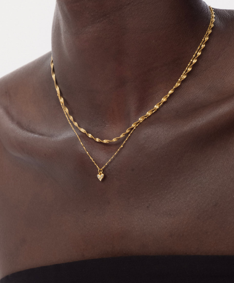 A close-up shot of a model wearing the gold Forever Necklace, featuring a delicate chain and a small heart pendant with sparkling stones, layered with a thicker twisted gold chain.