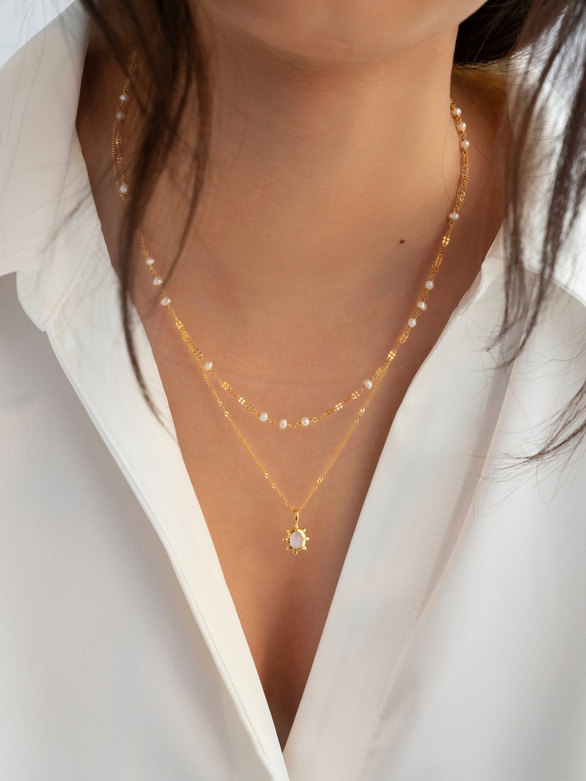 A close-up of a model wearing the gold Aurelia Necklace, featuring a delicate chain with small freshwater pearls, layered over a second gold necklace with a sun-shaped pendant.