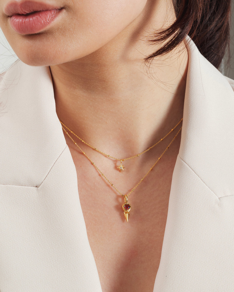 A close-up of a model wearing the Mrs H Spike Necklace in gold, featuring a pendant with a spike and a red garnet stone, layered with a shorter gold necklace with a star-shaped pendant.