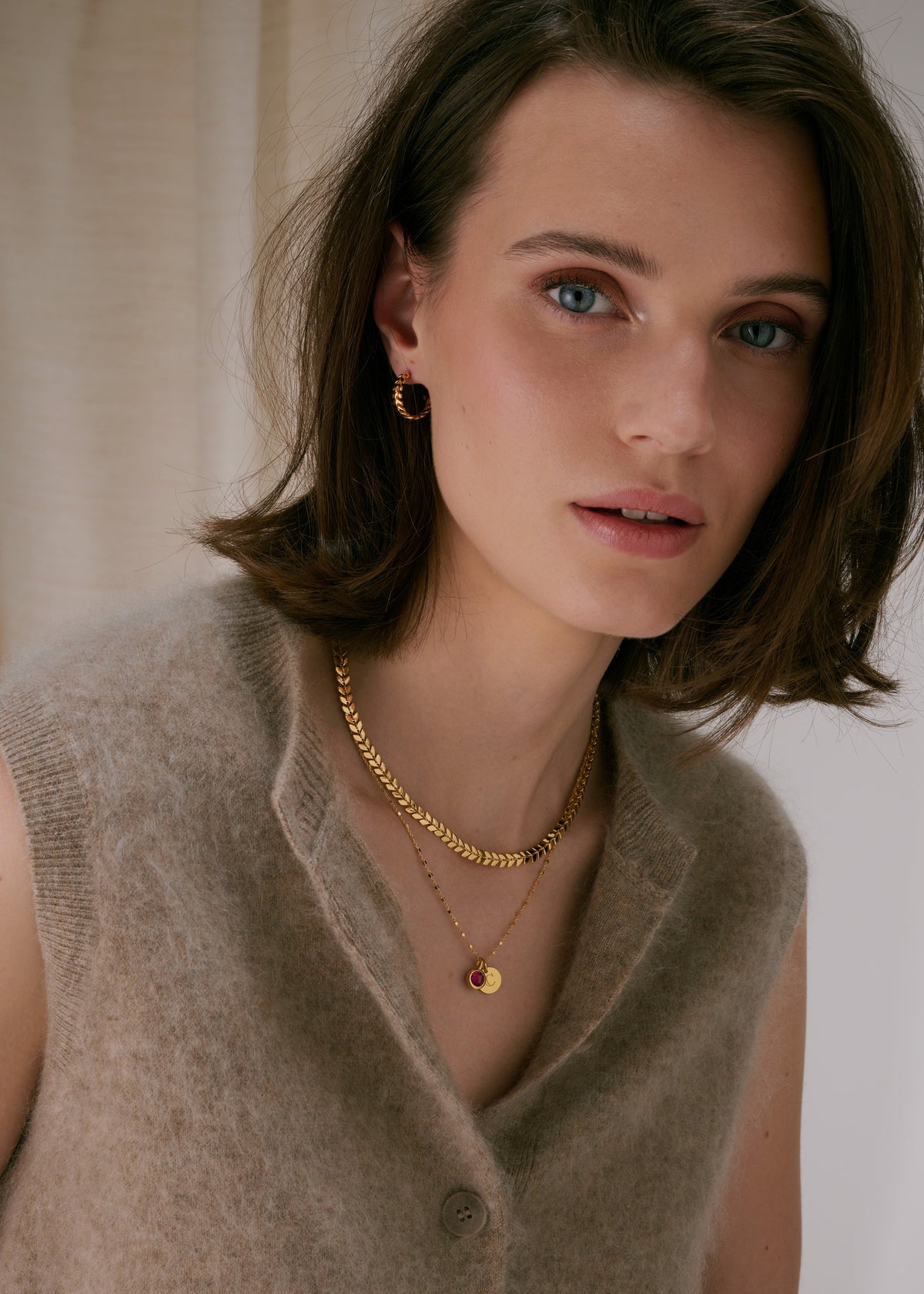 Layered gold necklaces featuring a leaf-patterned chain, a delicate chain, and a longer chain with a pink gemstone pendant, paired with twisted gold hoop earrings.