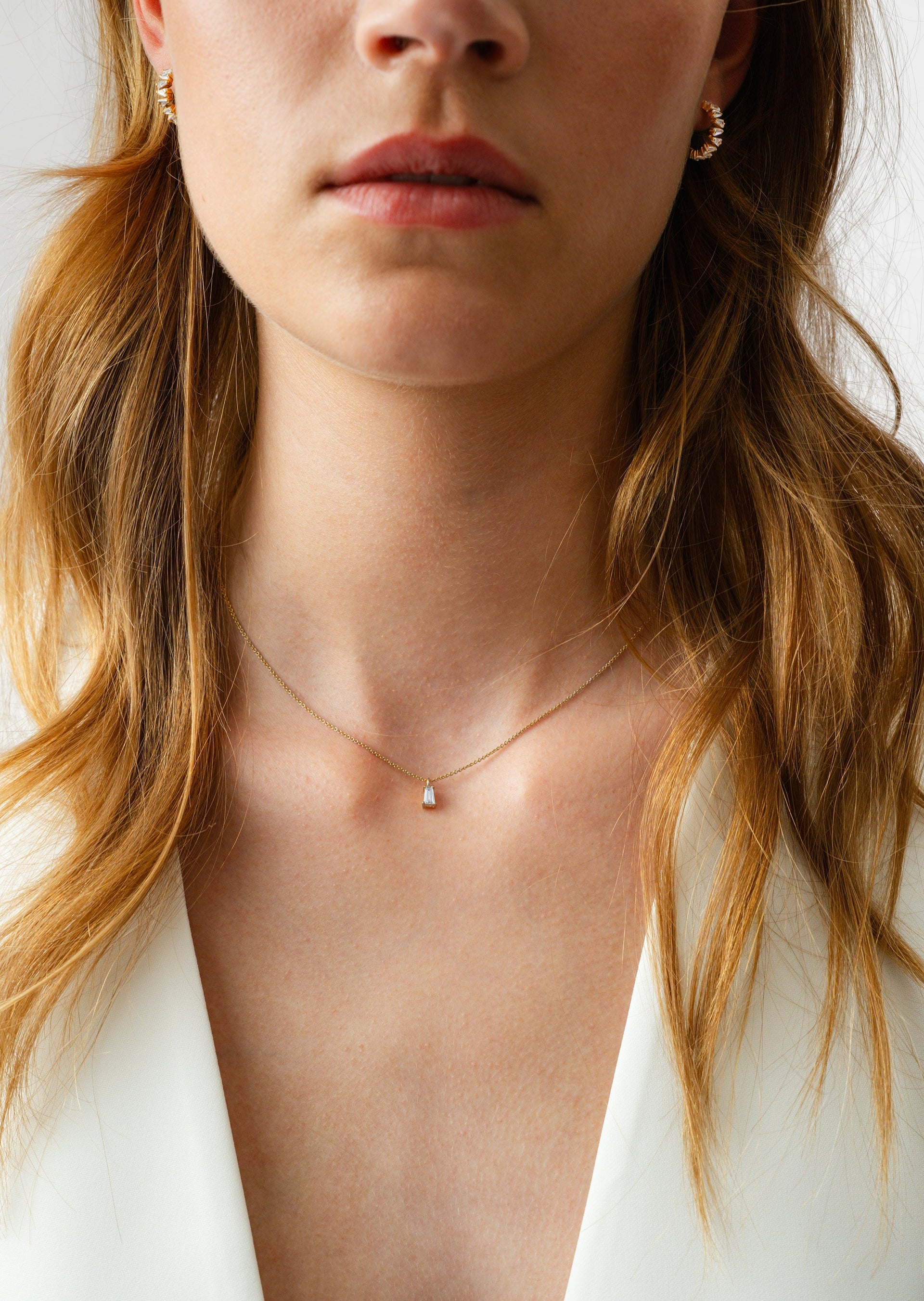A model wears the Iggy Necklace Gold, featuring a delicate gold chain and a single tapered baguette-cut stone pendant, styled with a pair of gold hoop earrings set with baguette stones.