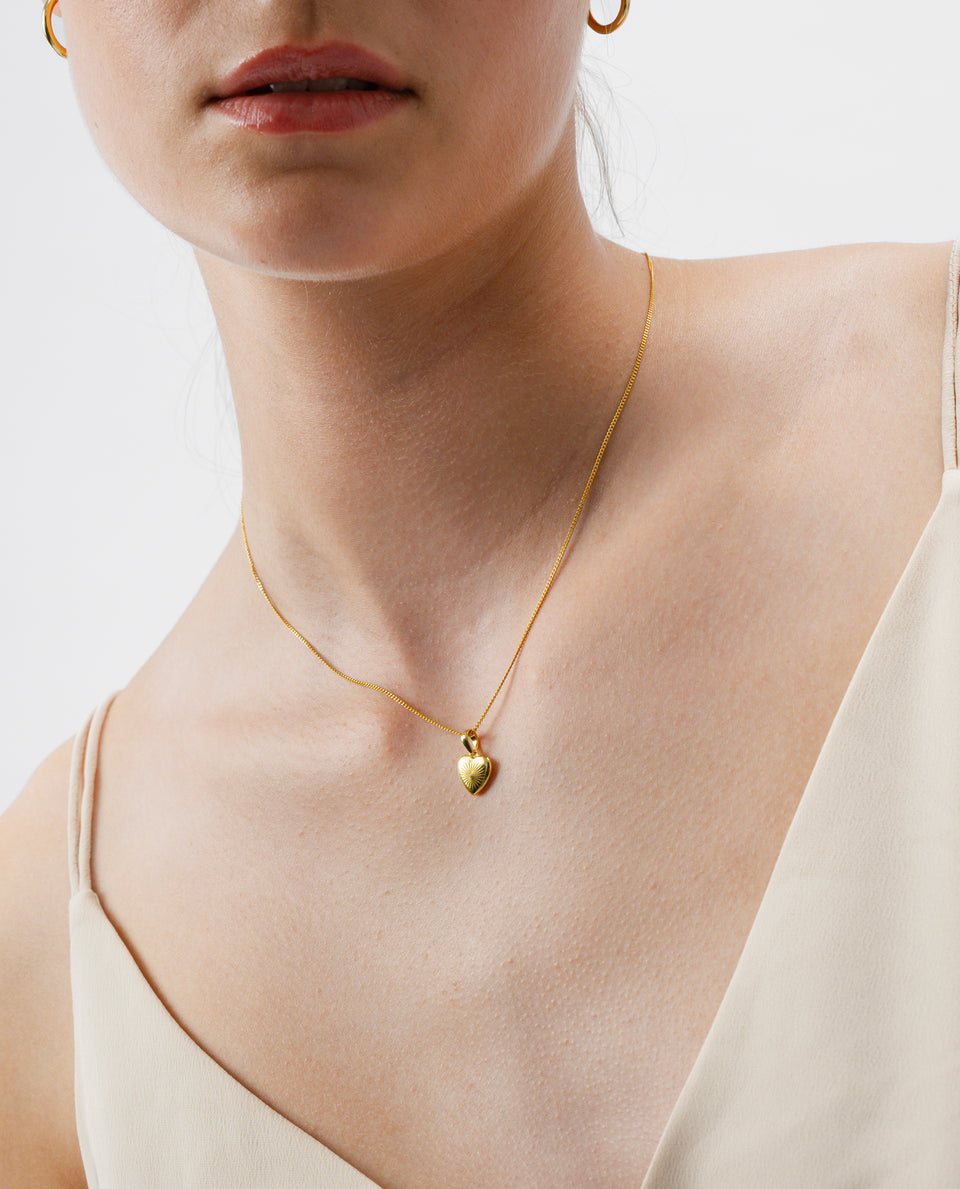 A close-up shot of a model wearing the Sunburst Heart Necklace in gold, which features a fine chain and a heart-shaped pendant with a sunburst pattern. A small gold hoop earring is also visible.