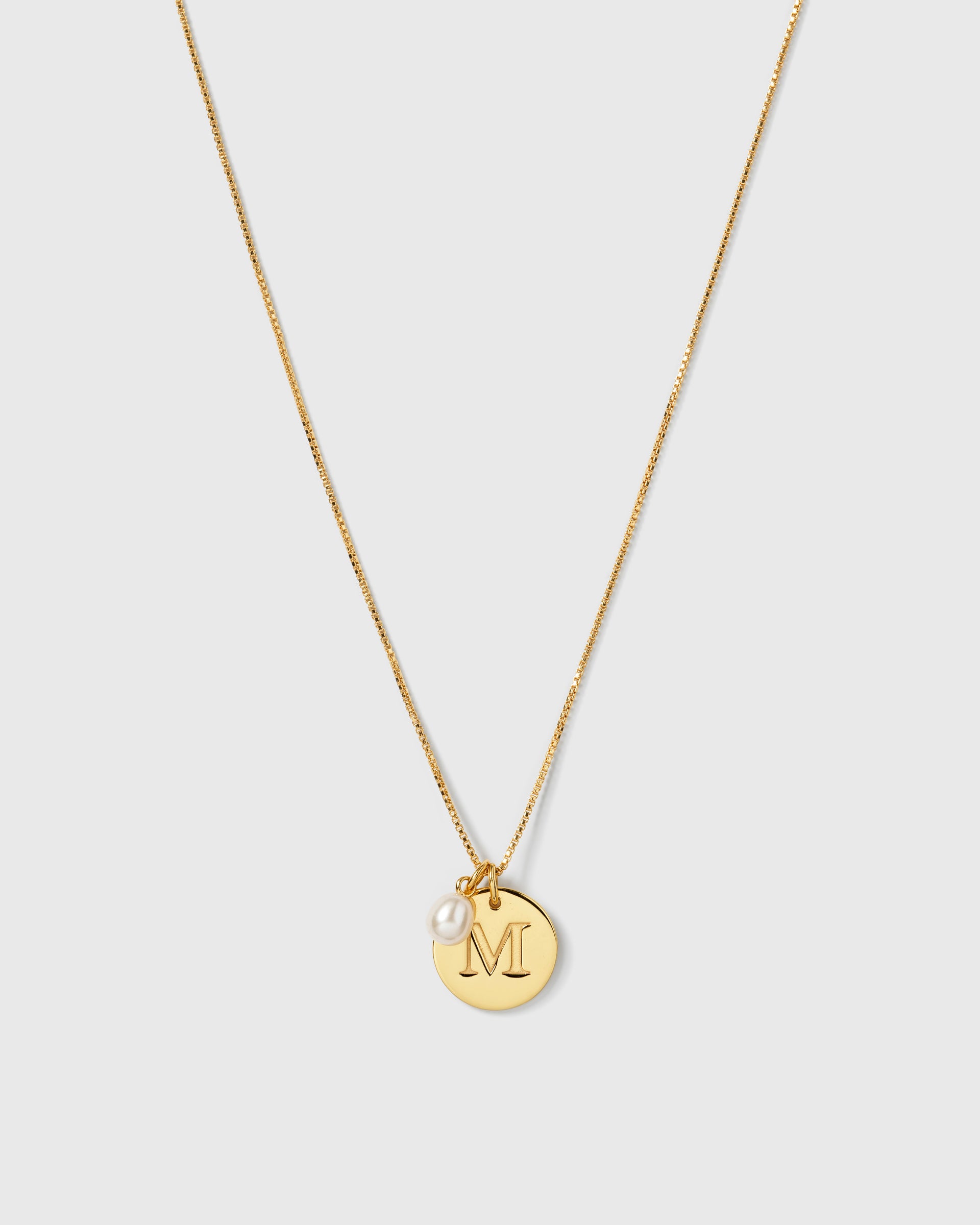 A gold necklace with a fine box chain, featuring the Beloved Pearl Pendant and a circular gold pendant engraved with the letter 'M'.