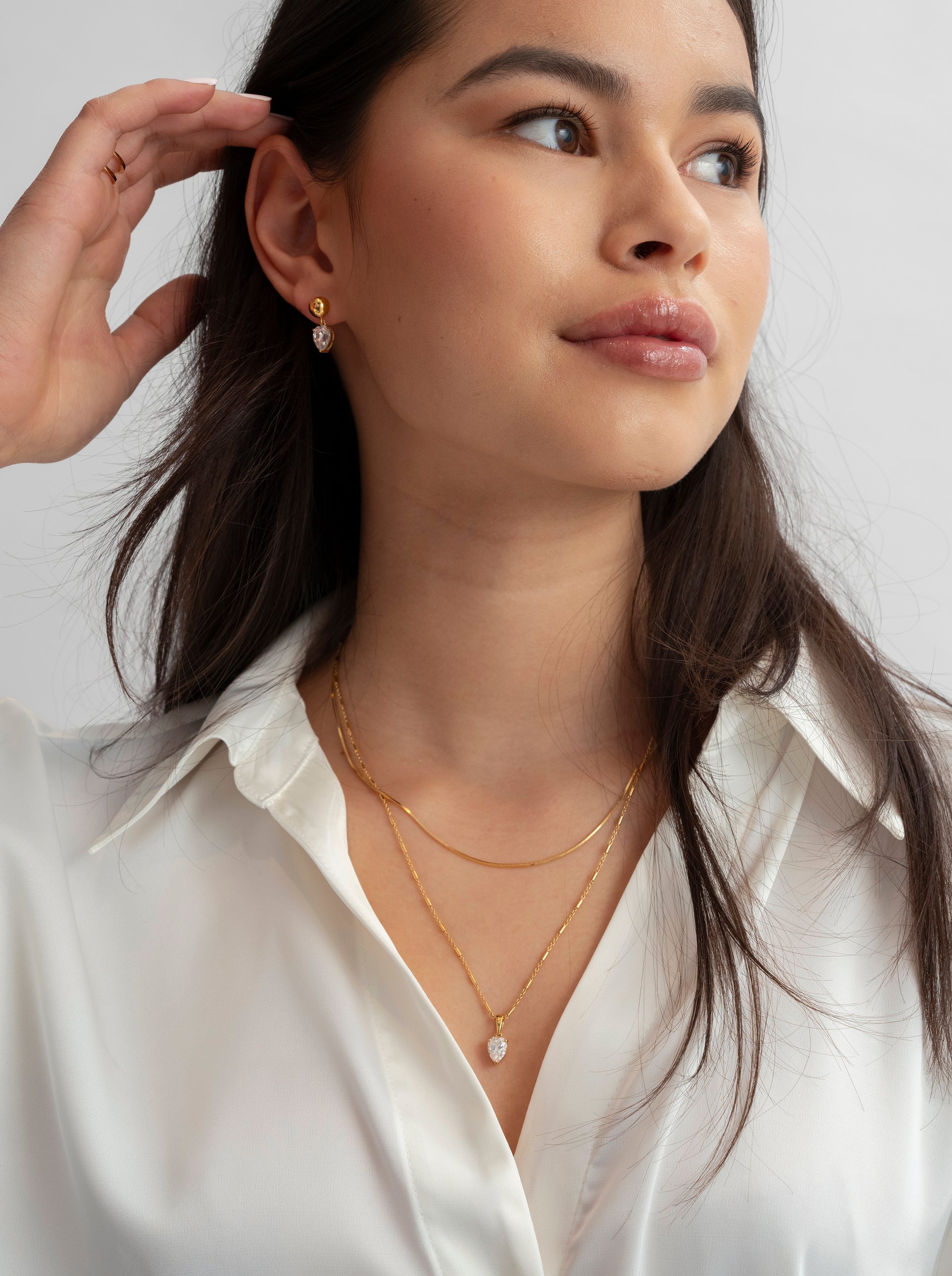 Layered gold necklaces with a drop-shaped clear stone pendant, worn with matching earrings and a gold double-band ring.