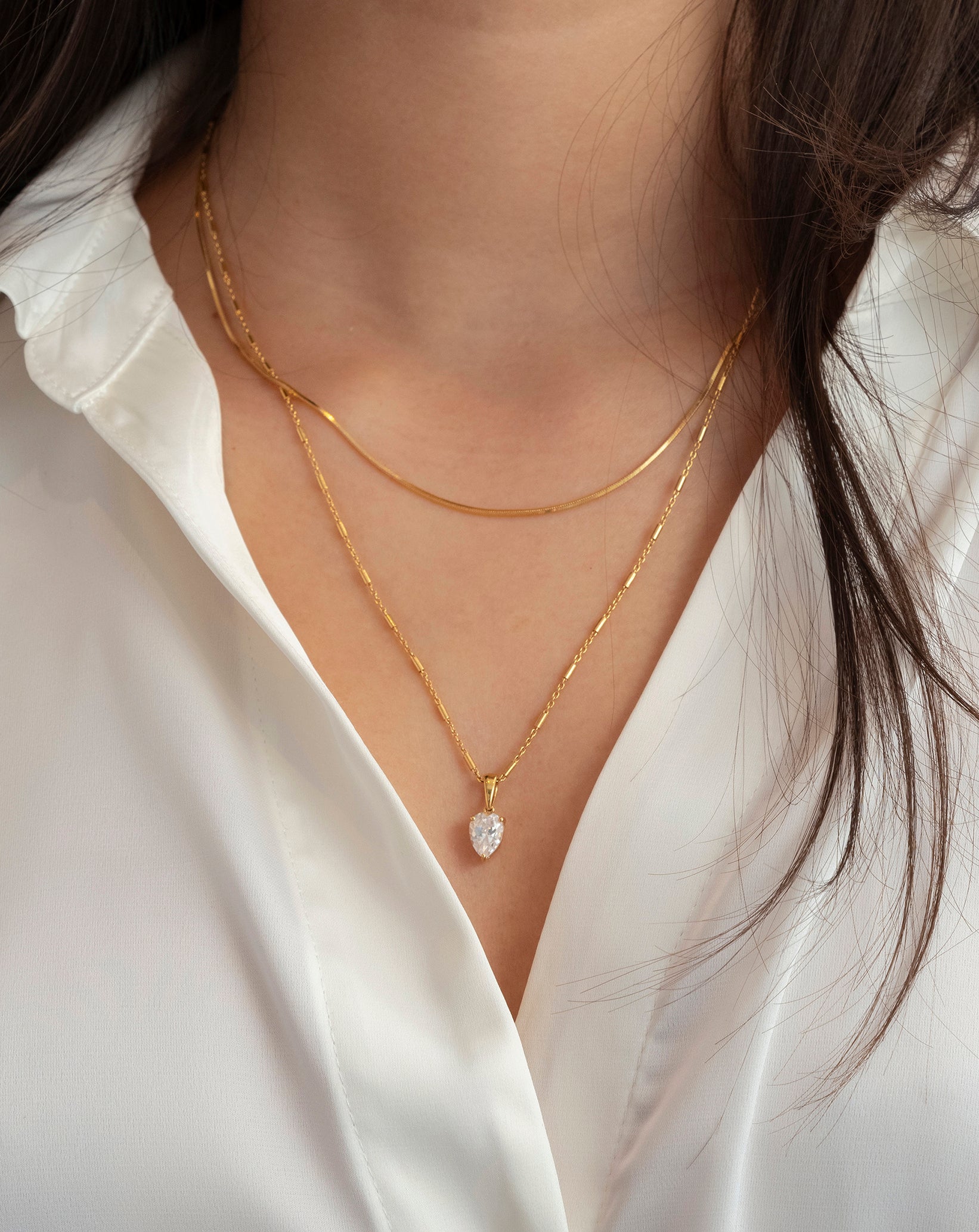 A close-up shot of a model wearing the Katie Necklace in gold, with its drop-shaped cubic zirconia pendant, layered with a shorter gold snake chain necklace.