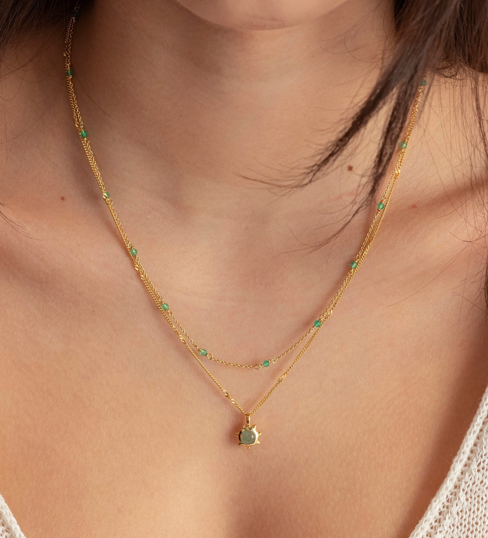 A close-up shot of a model wearing the Florentina Necklace in gold, which features a flower-shaped green jade pendant on a twisted chain, layered with another gold necklace adorned with small green jade beads.
