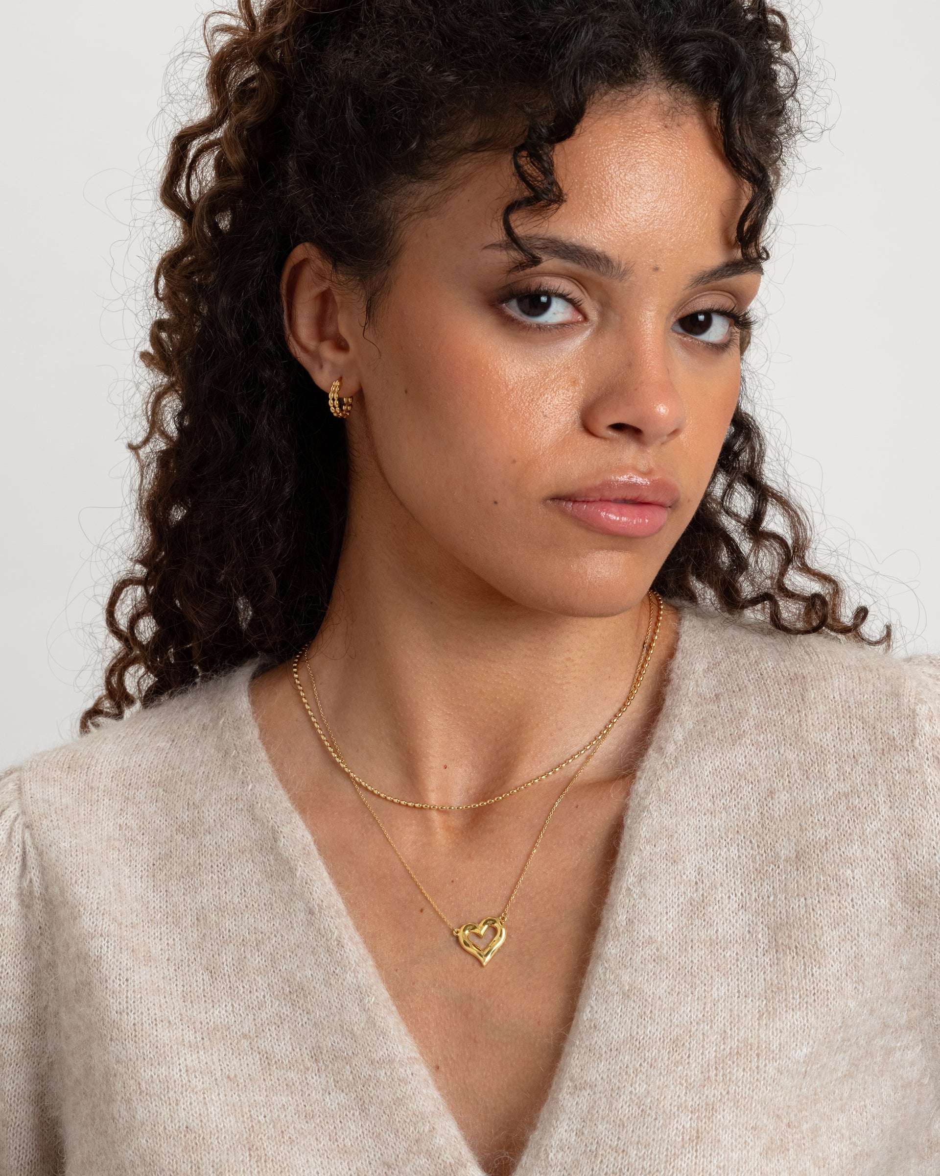 A model showcasing the gold Puff Heart Necklace, featuring its chunky heart-shaped pendant, layered with a shorter delicate gold chain, and paired with small textured gold hoop earrings.
