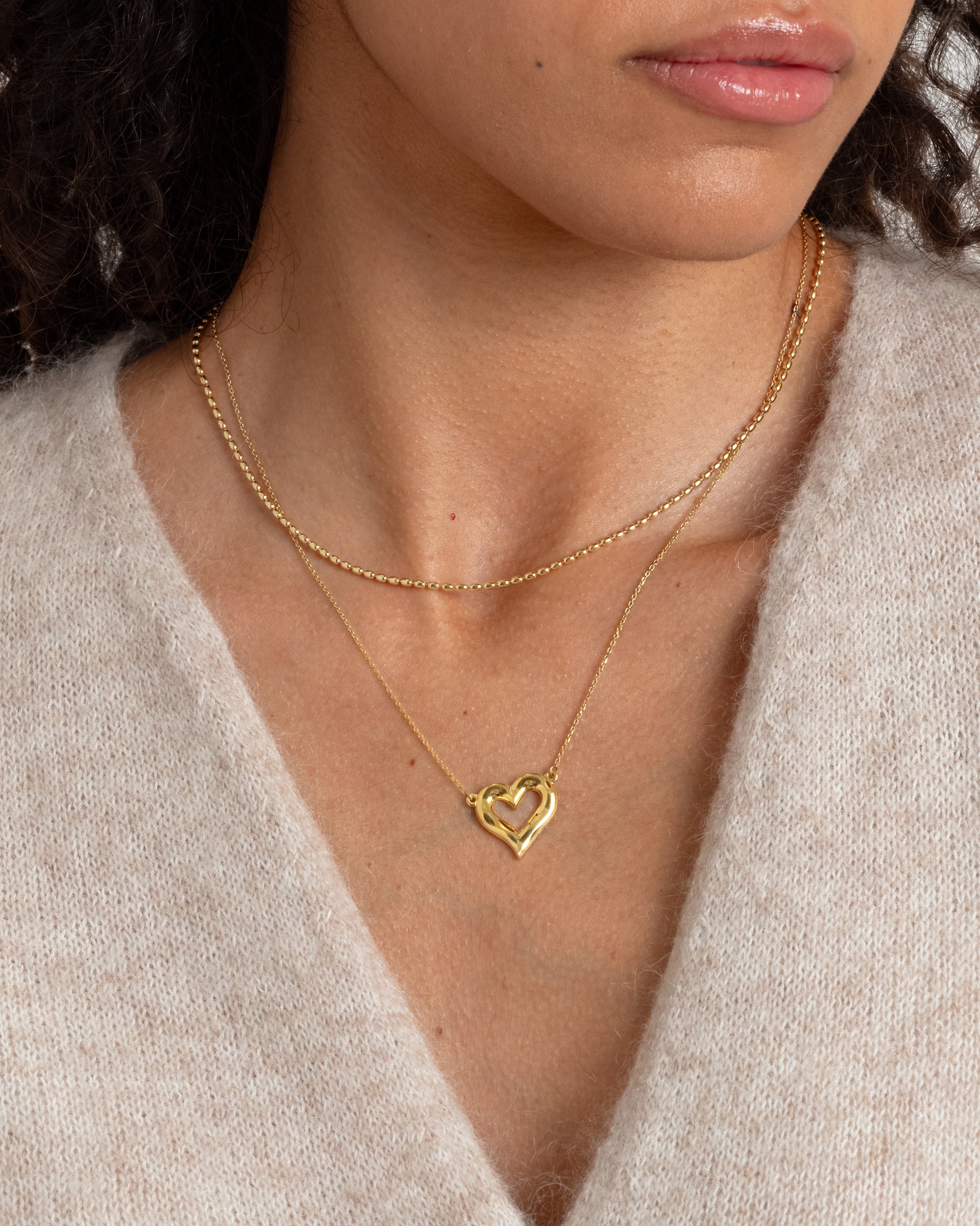 A close-up shot of a model wearing the Puff Heart Necklace in gold, featuring its chunky heart-shaped pendant, layered with a shorter delicate beaded gold necklace.