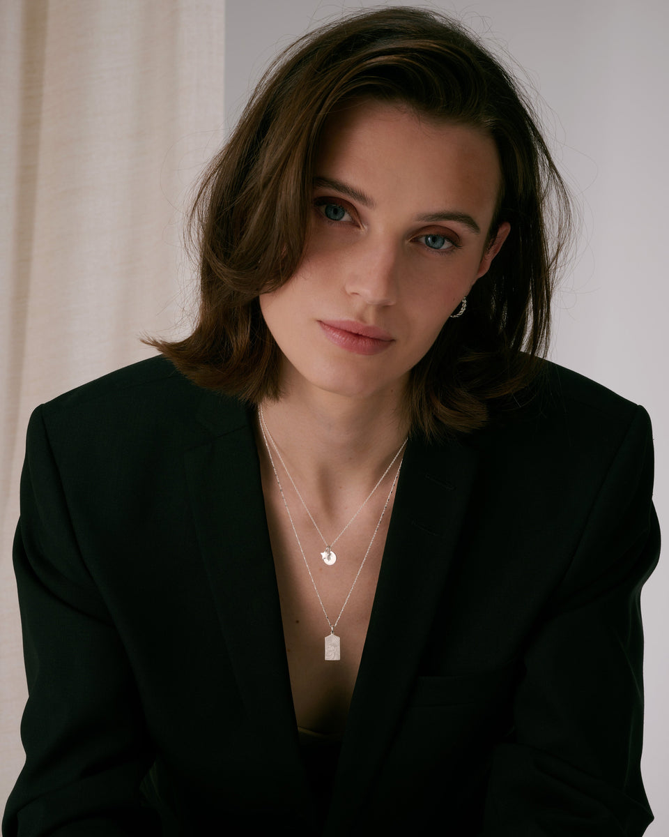 Three layered silver necklaces with letter pendant, star charm, and rectangular pendant on fine chains, paired with small silver hoop earrings.