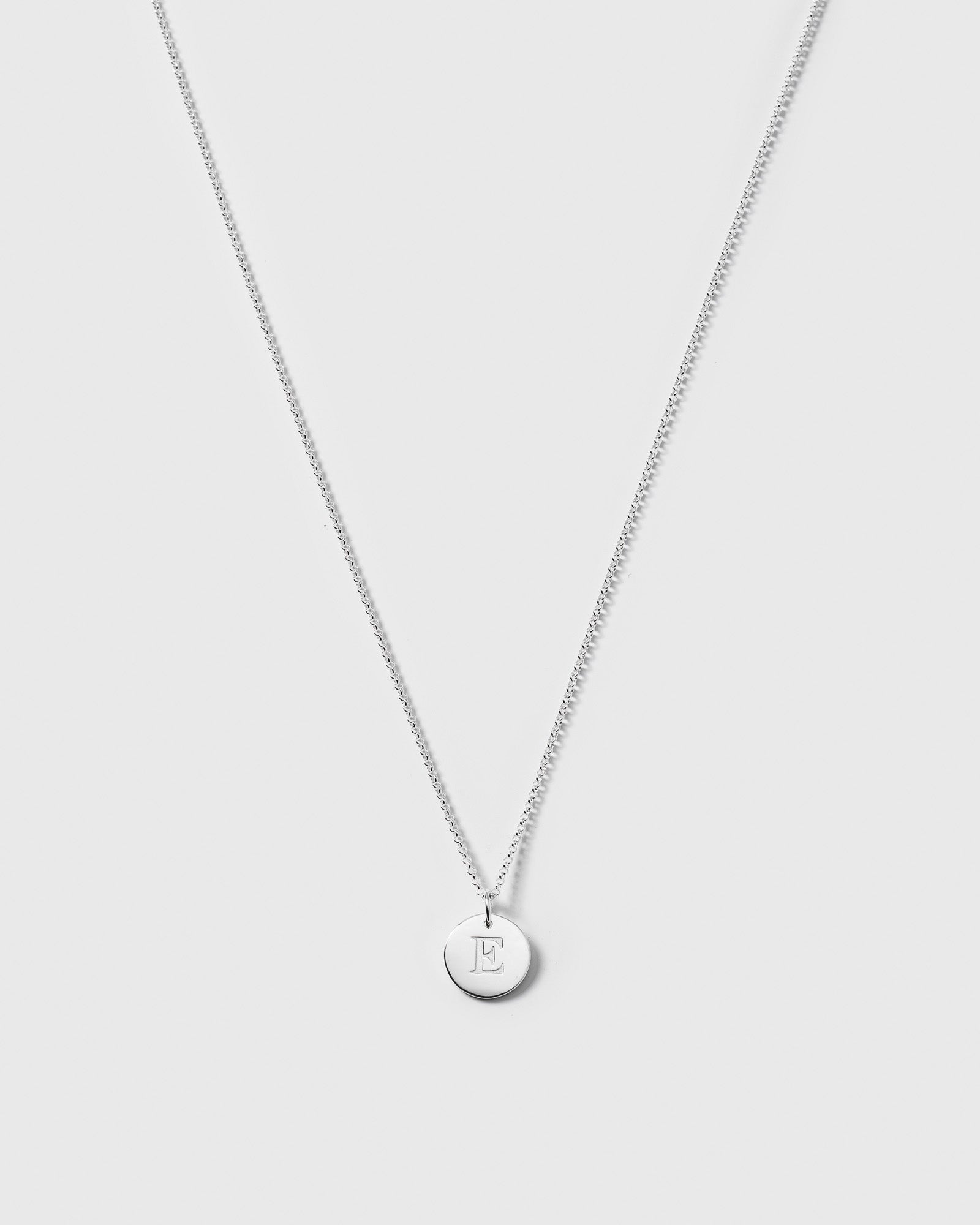 A product shot of the Beloved Medium Rolo Chain in silver, shown styled with a matching round silver pendant engraved with the letter E.
