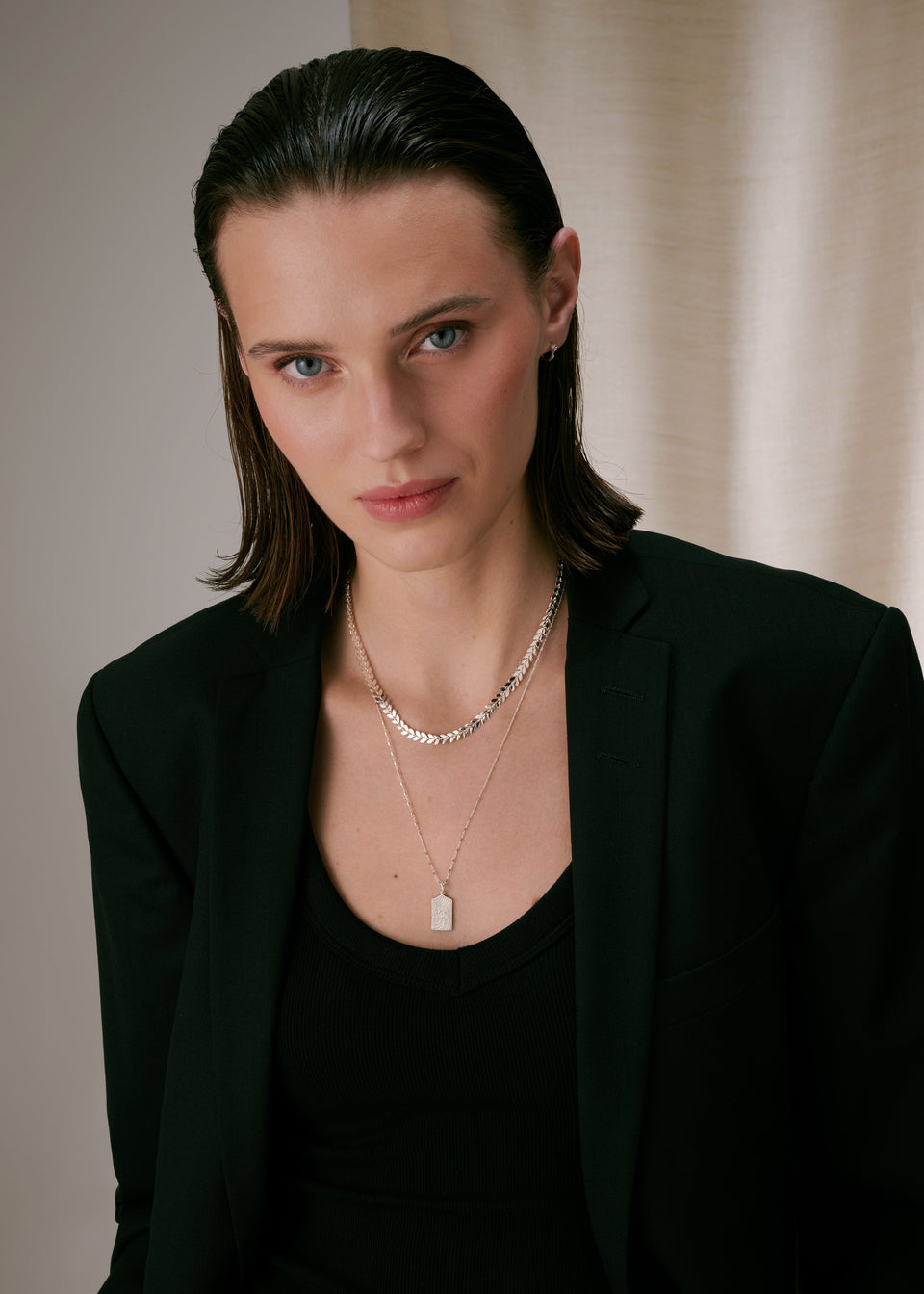 A short silver necklace with a leaf-like pattern, layered with a longer delicate silver chain featuring a rectangular pendant, paired with a small silver hoop earring.