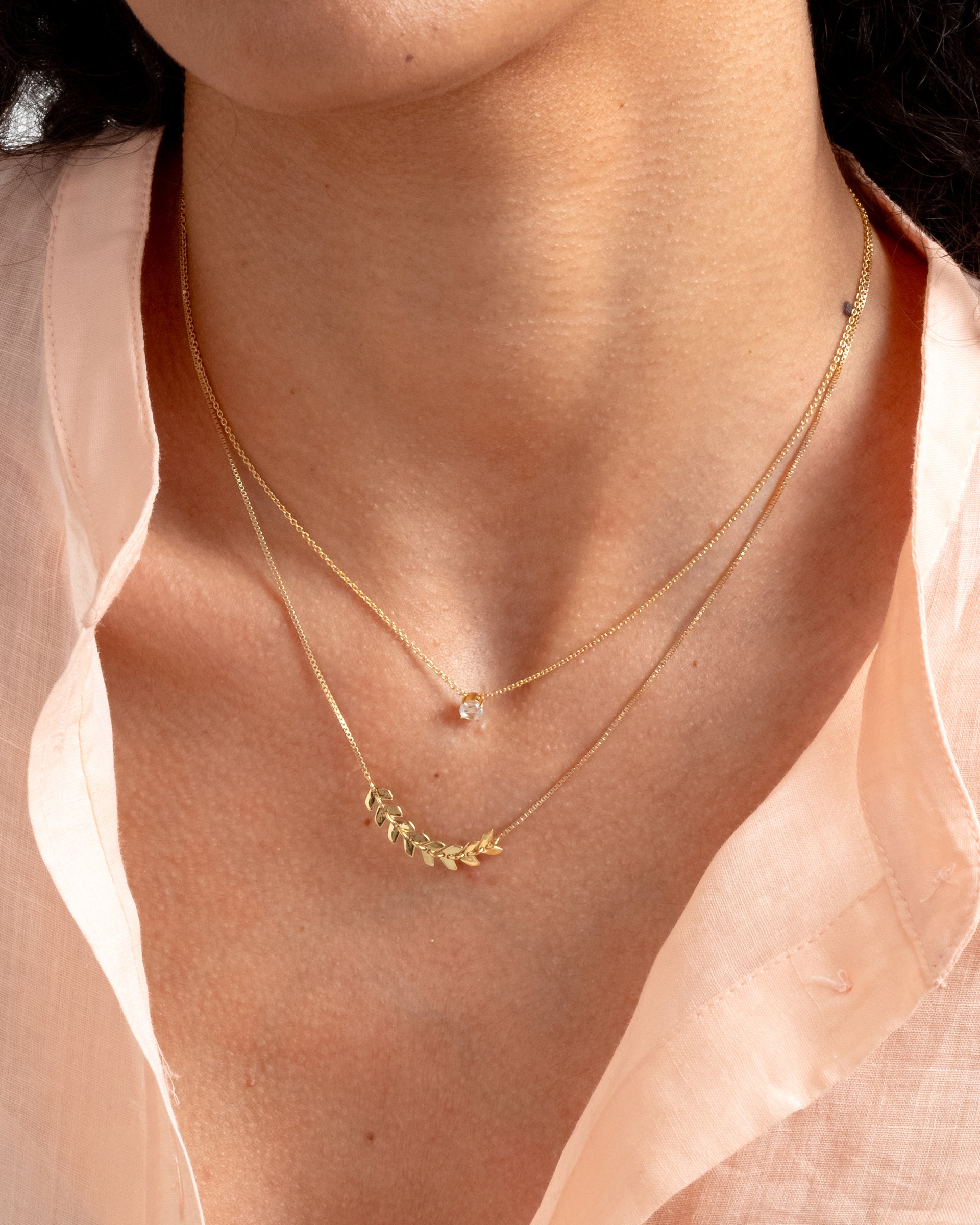 A close-up shot of a model wearing the Layers Simone Necklace in gold, featuring a delicate leaf pattern, layered with a thin gold solitaire necklace.