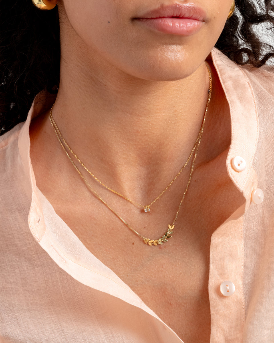 A model wears the gold Layers Simone necklace with a delicate leaf pattern, layered with a shorter gold necklace with a small crystal pendant.