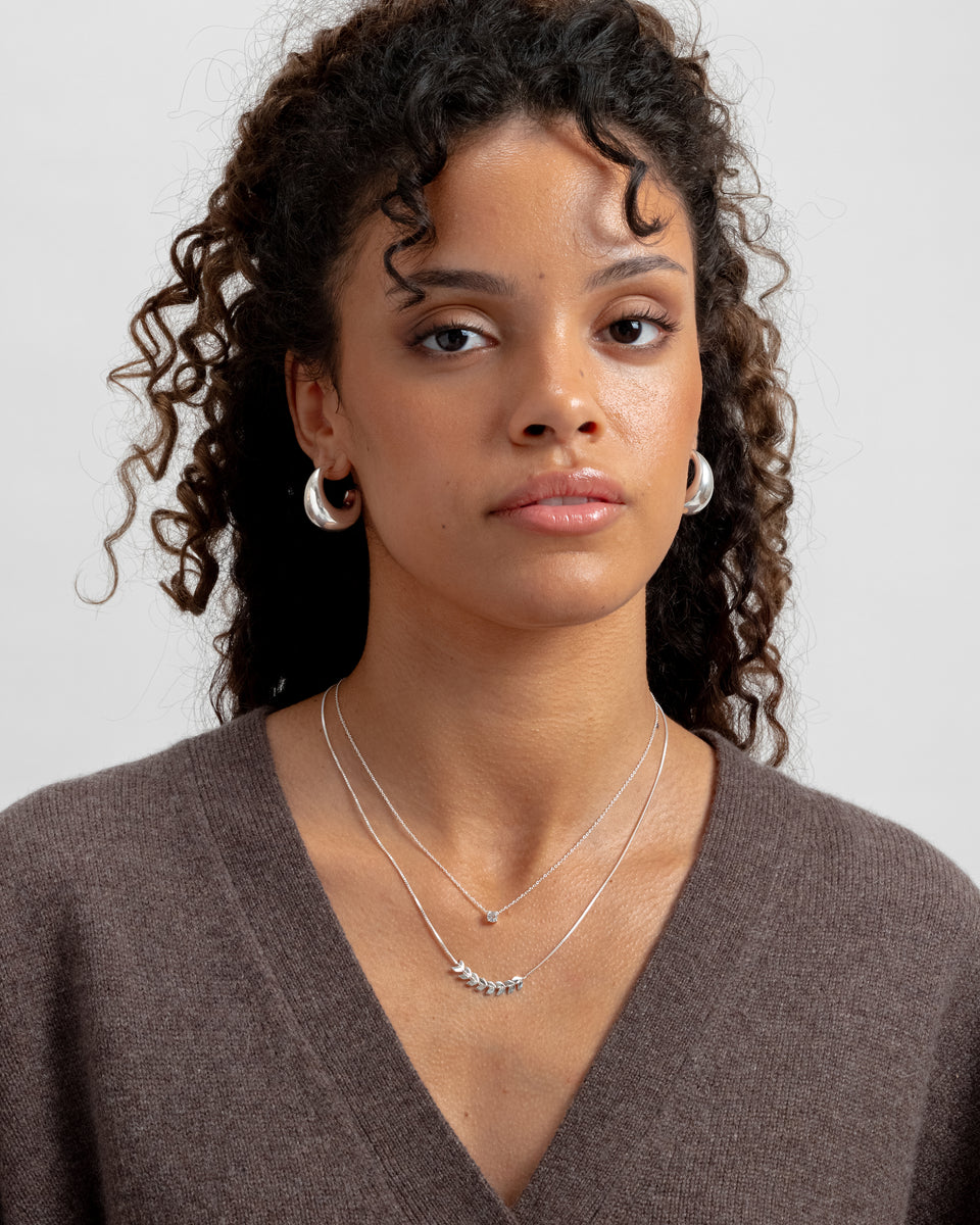 Layered silver necklace with delicate leaf-like details on the longer chain, paired with a shorter fine chain necklace, and chunky silver hoop earrings.