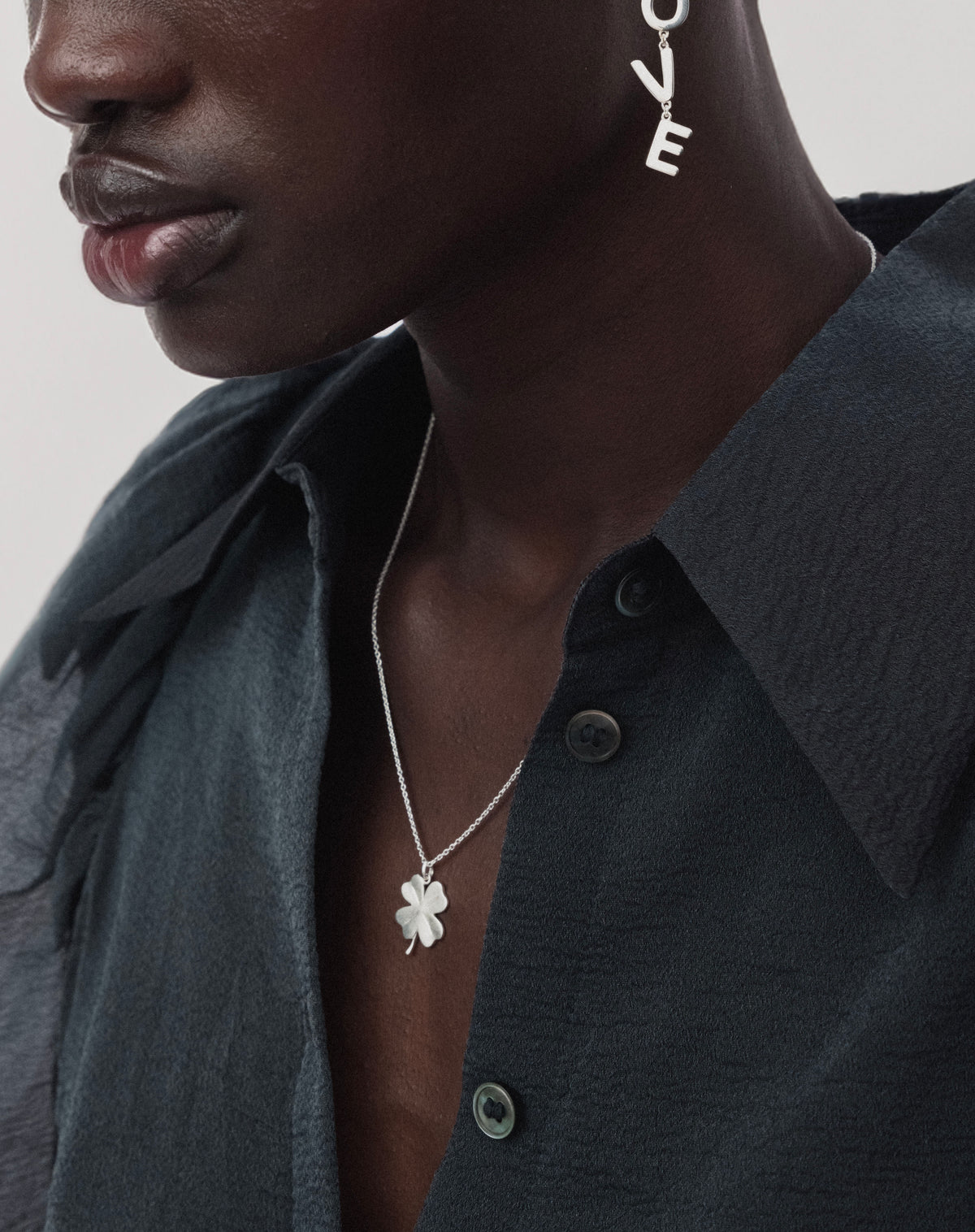 A close-up of a model wearing the Beaches Fortune Necklace in silver, featuring a fine chain and a matte four-leaf clover pendant. She is also wearing a silver drop earring that spells out the word love.
