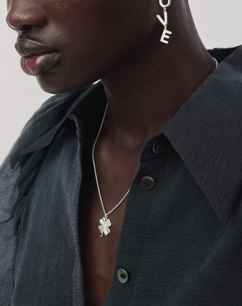 A close-up of a model wearing the Beaches Fortune Necklace in silver, featuring a fine chain and a matte four-leaf clover pendant. She is also wearing a silver drop earring that spells out the word love.