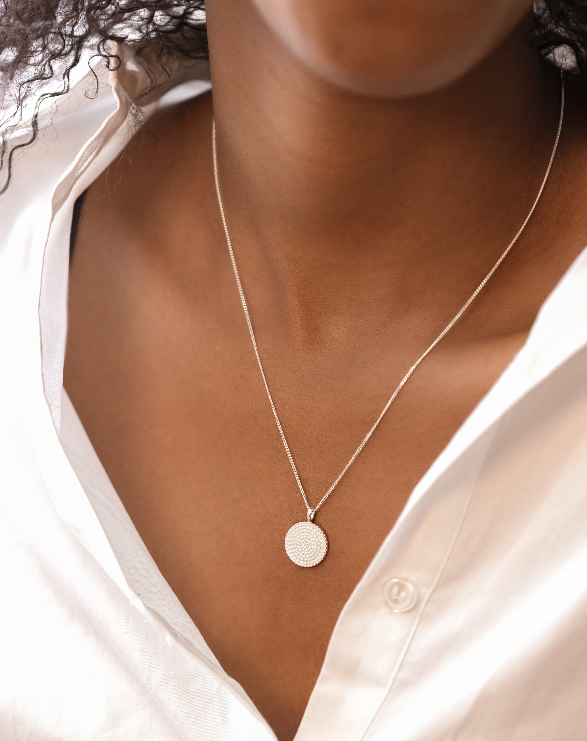 A close-up on a model wearing the Lucky Coin Heal Necklace in silver, styled with a white shirt. The image highlights the delicate chain and the round pendant with its textured sun pattern.
