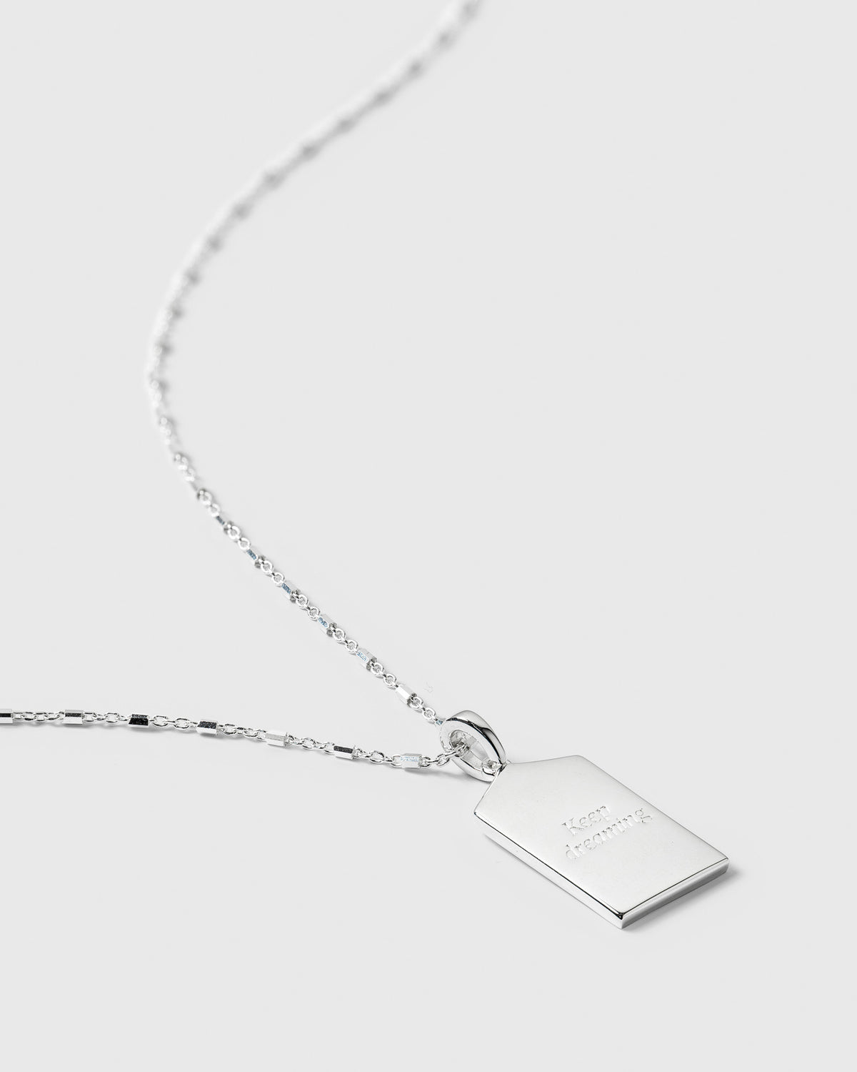 A close-up of the silver Lucky Coin Keep Dreaming Necklace, showing the back of the rectangular pendant engraved with the words 'Keep dreaming'.