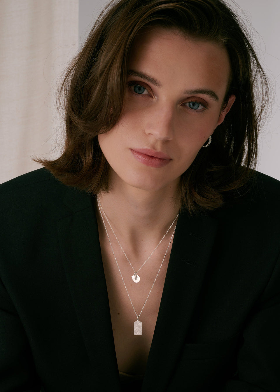 A silver coin pendant necklace layered with a longer silver necklace featuring a rectangular pendant, paired with a small silver hoop earring.