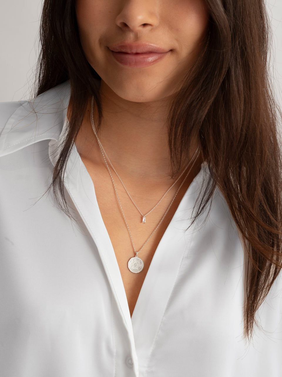 A close-up of a model wearing the silver Lucky Coin Navigate Necklace, featuring a round compass pendant, layered with a shorter silver necklace with a small rectangular charm.