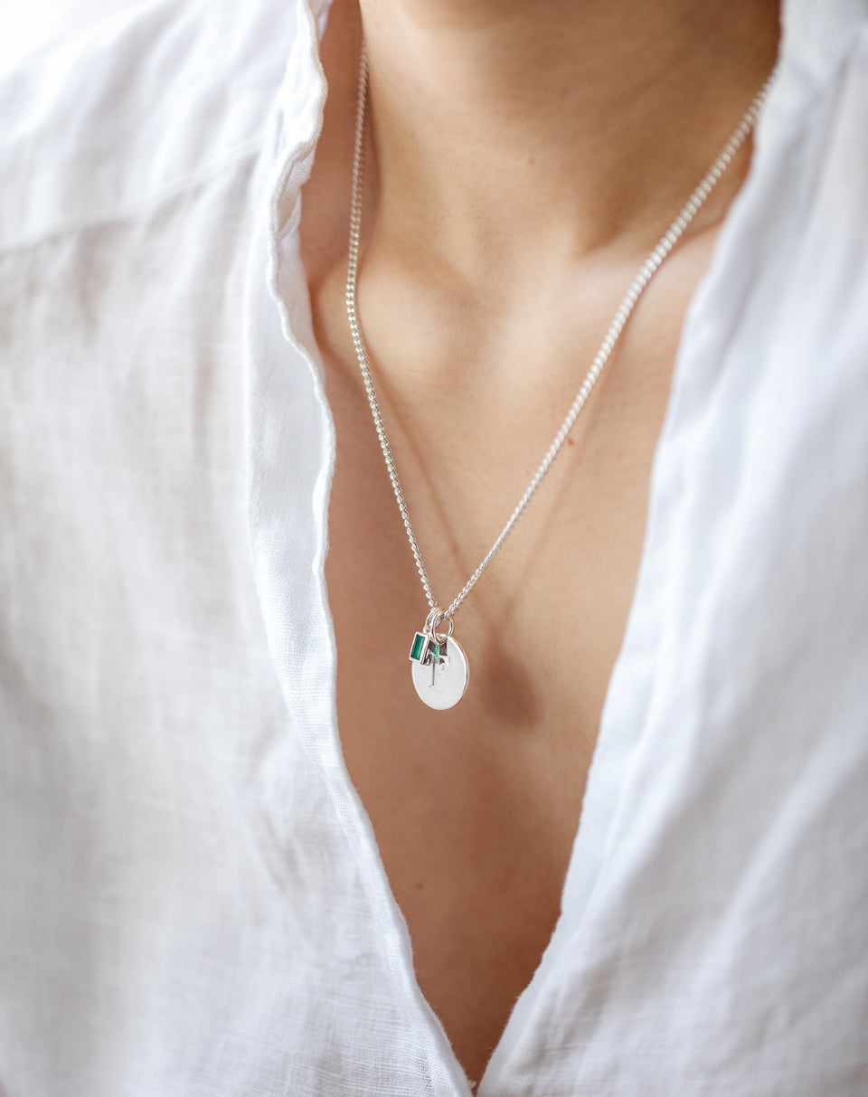 A close-up shot of a model wearing the Beloved Squared Pendant in silver with a green onyx, styled on a silver chain alongside a round, engraved letter pendant.