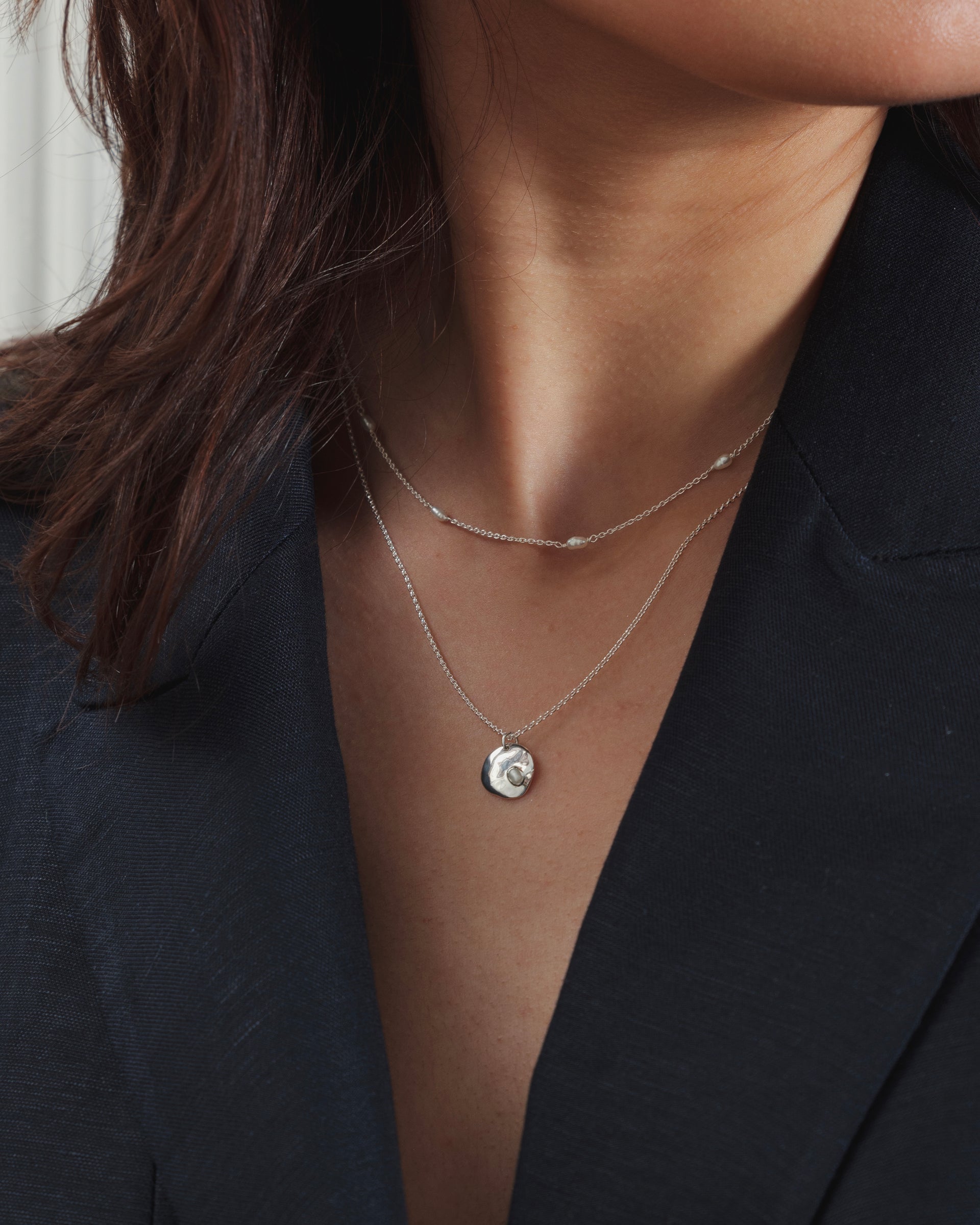 A model wears the Syster P Treasure Multi Pearl Necklace in silver, featuring a delicate chain with small freshwater pearls, layered with a second silver necklace that has an organic-shaped pendant with an embedded pearl.